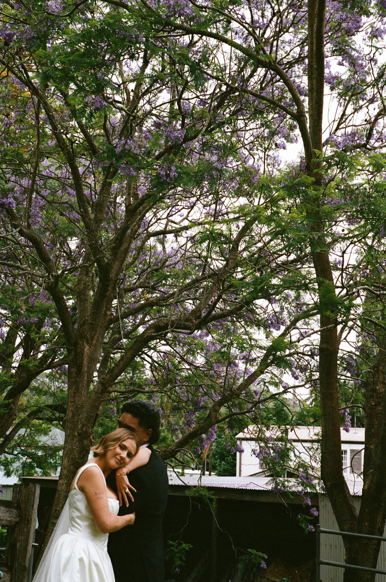 A white wedding dress flows beneath blooming purple jacaranda trees in a romantic garden setting.