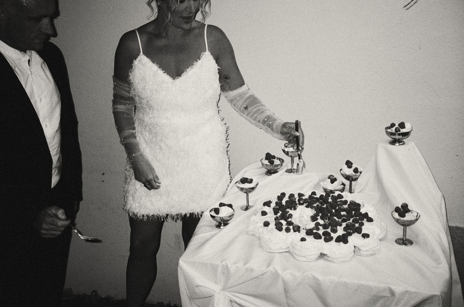 Black and white photo showing a table covered in white cloth with scattered dark berries and small desserts.