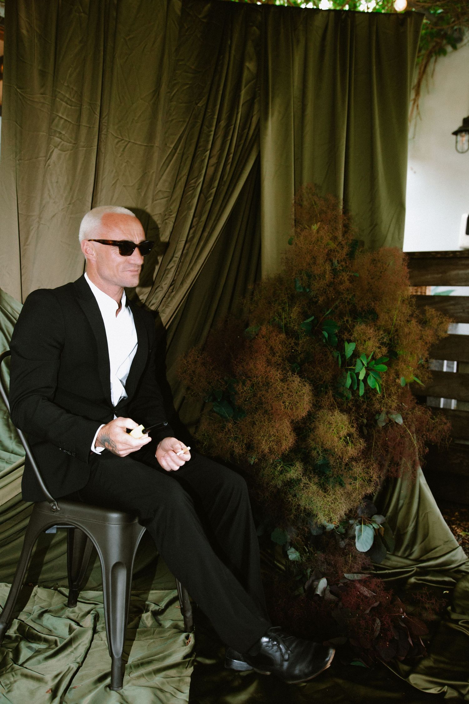 A formally dressed man in sunglasses sitting on a black chair against a dark green backdrop.