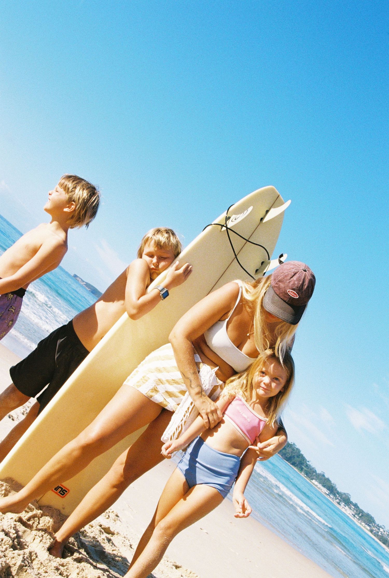 Family of four with surfboards on a sunny beach, two kids and adults in swimwear enjoying a summer surf day.