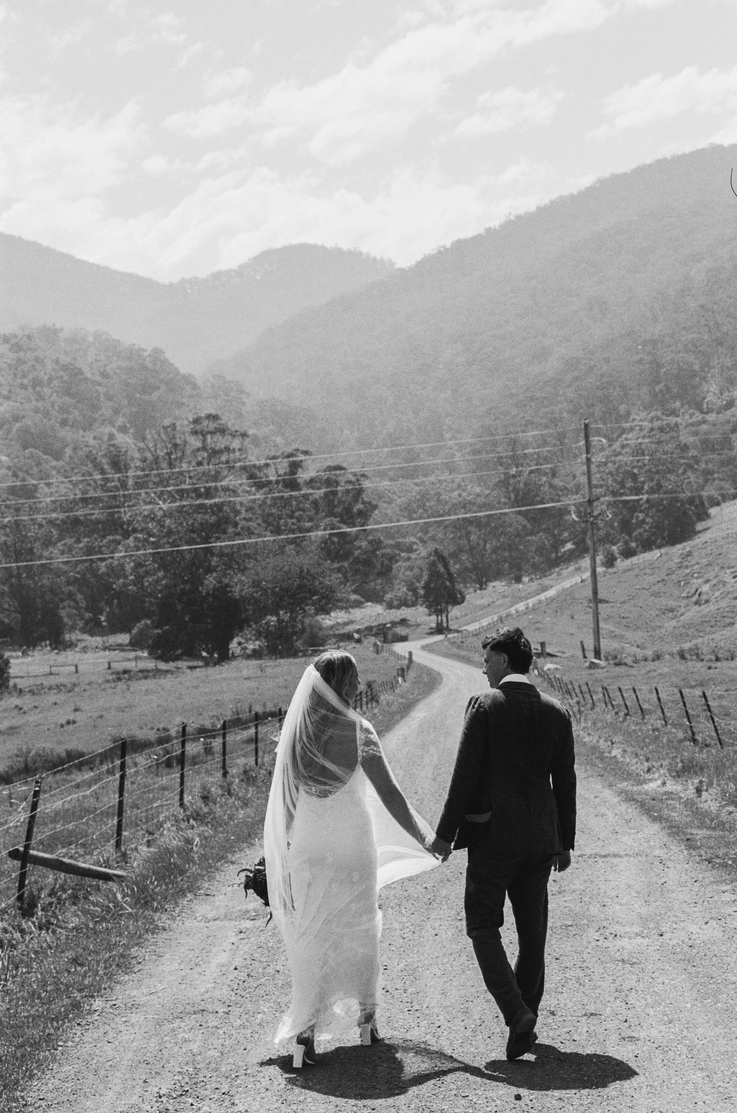 Newlyweds walking hand in hand down a country road with mountains in the background.