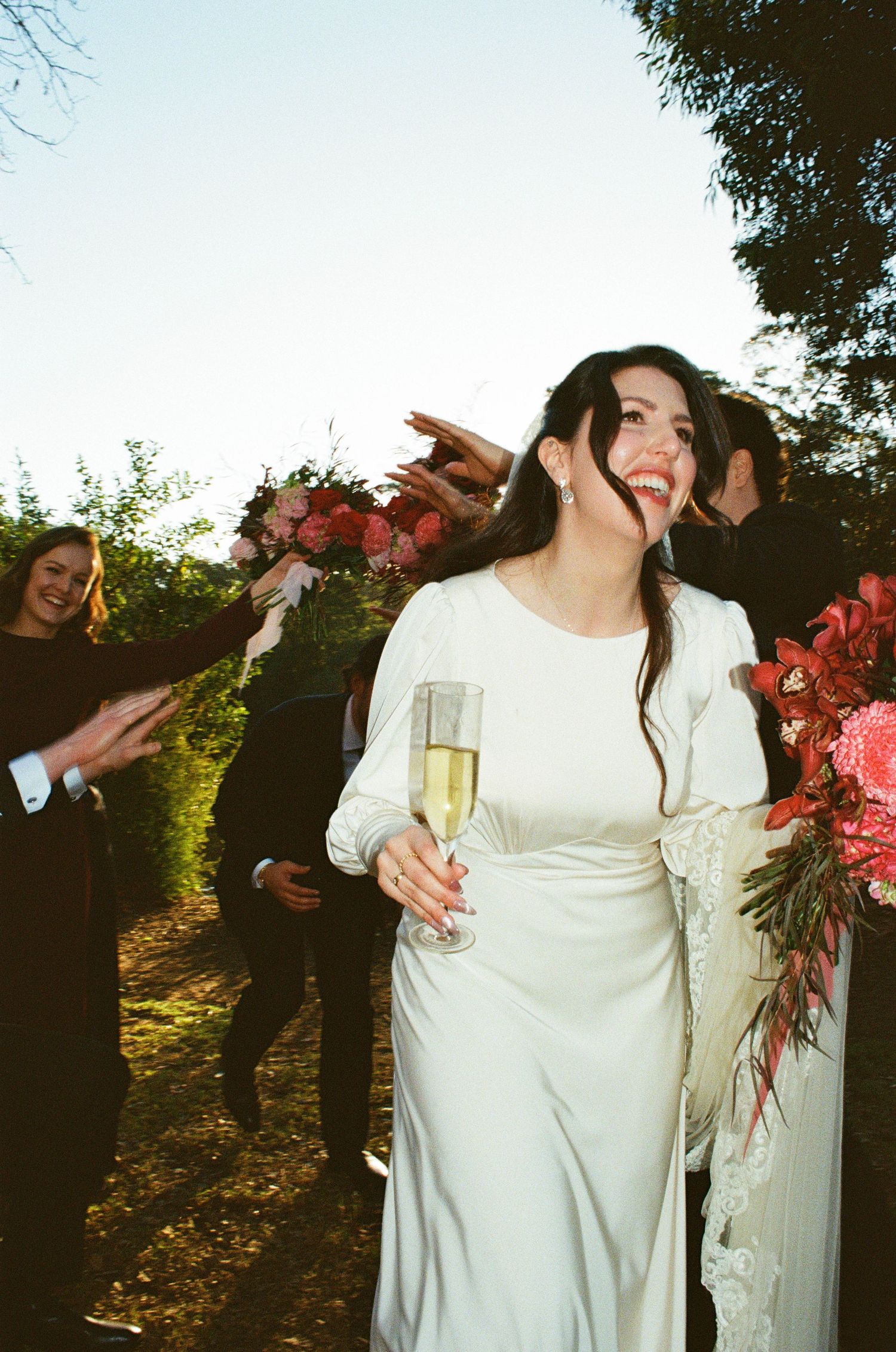 A bride in a white long-sleeve dress laughs joyfully while holding a champagne glass during an outdoor wedding celebration.