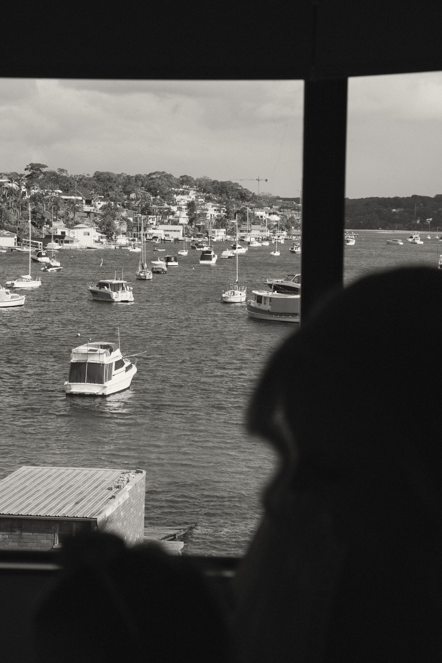A silhouetted figure overlooks a scenic harbor filled with moored boats and yachts on a cloudy day.