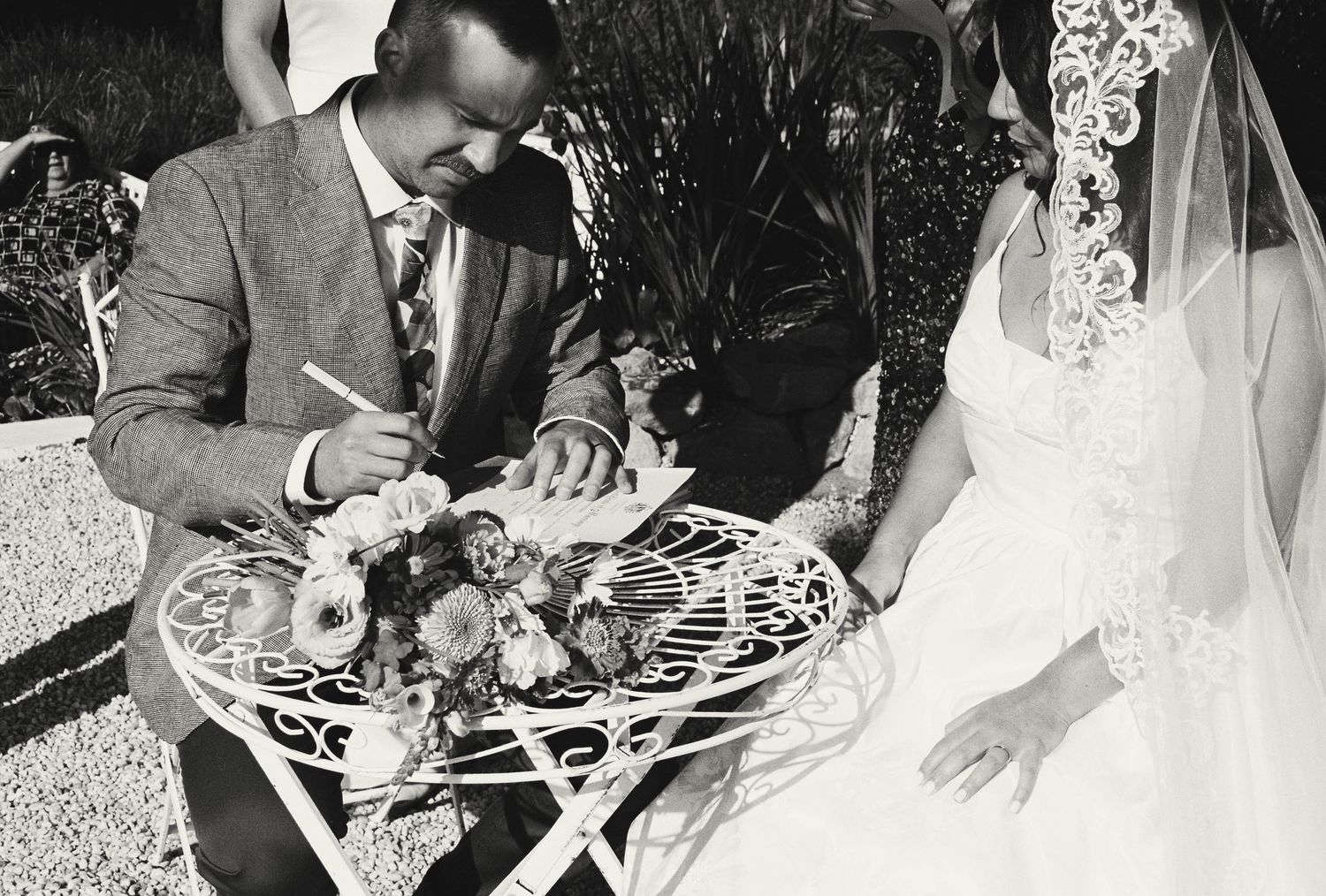 Newlyweds signing their marriage certificate at a white decorative table during an outdoor ceremony.