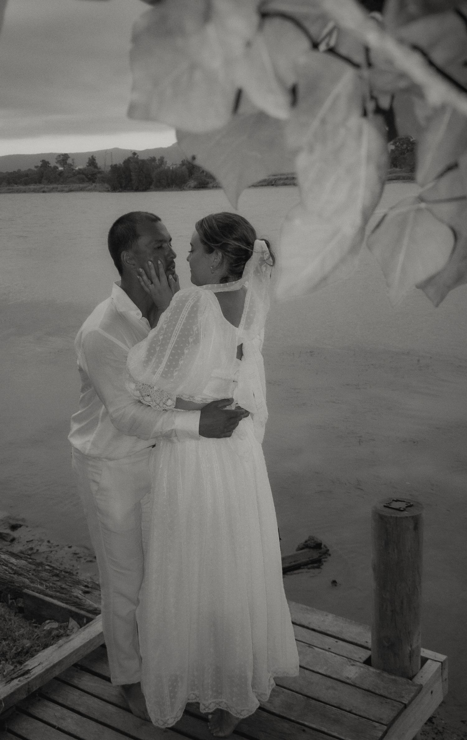 Black and white photo of a couple embracing on a coastal dock in wedding attire.