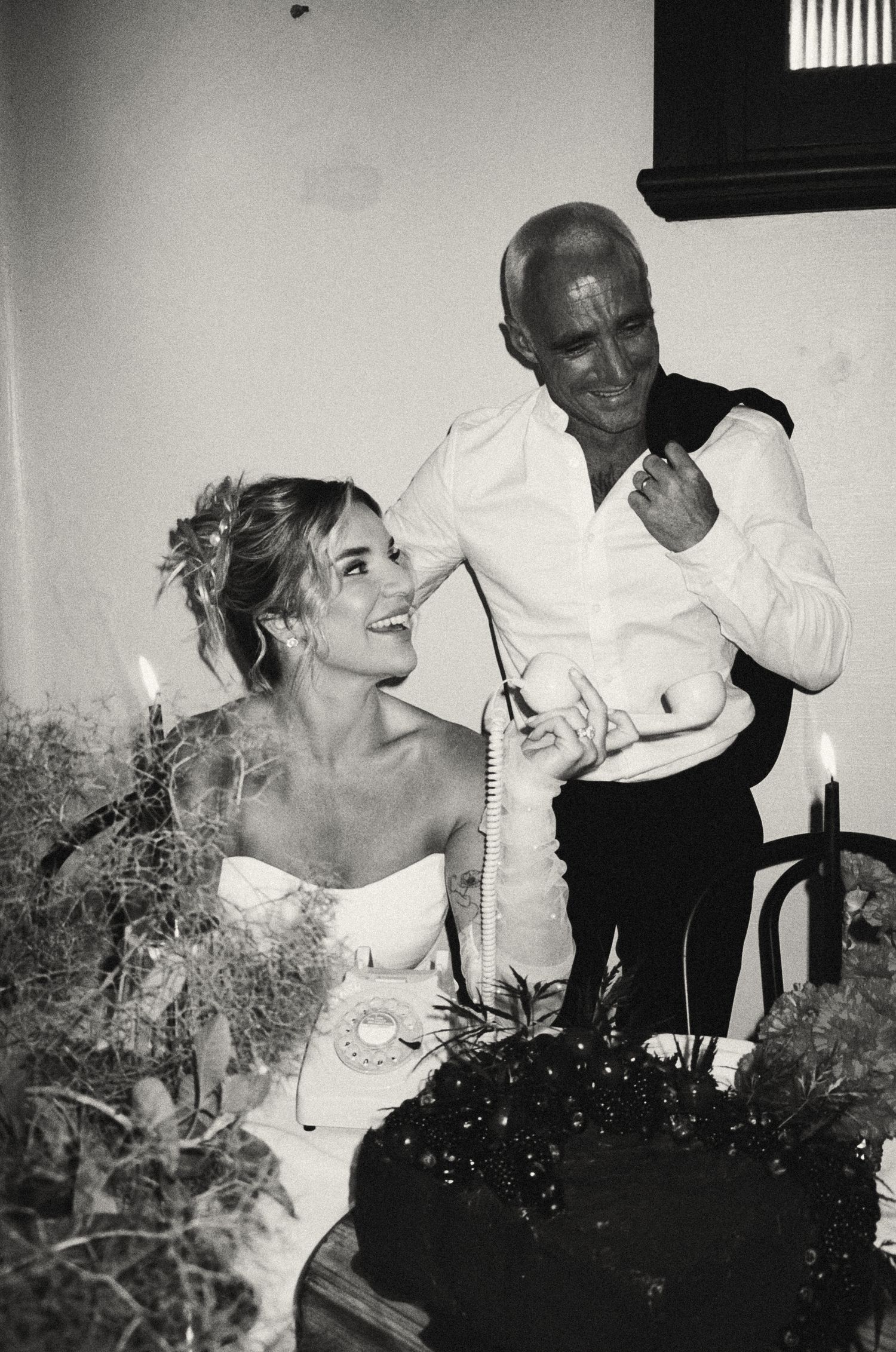 A black and white candid photo captures a joyful moment between two people at a wedding celebration.