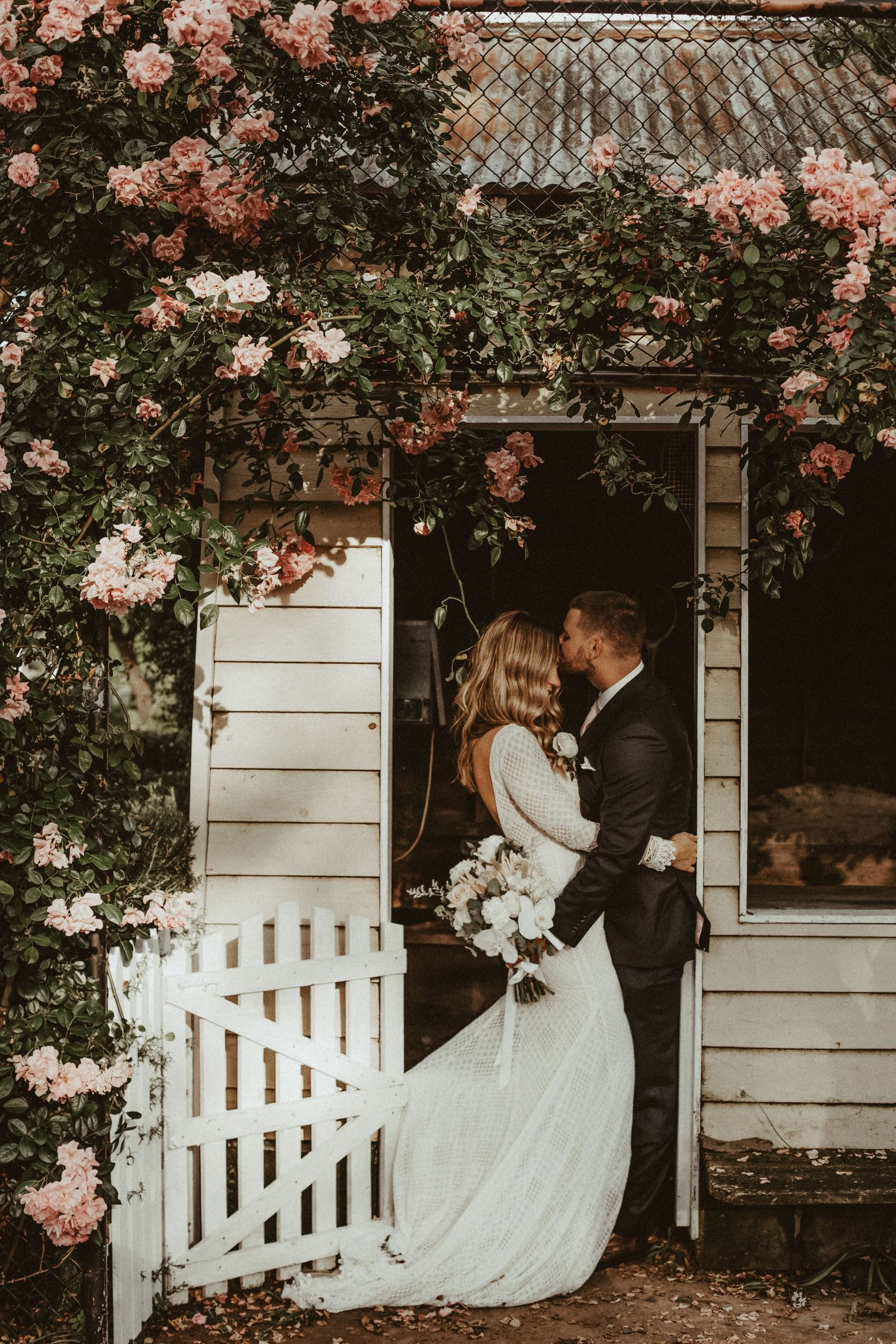 Romantic couple embraces in doorway of rustic white cottage surrounded by climbing pink roses and greenery.