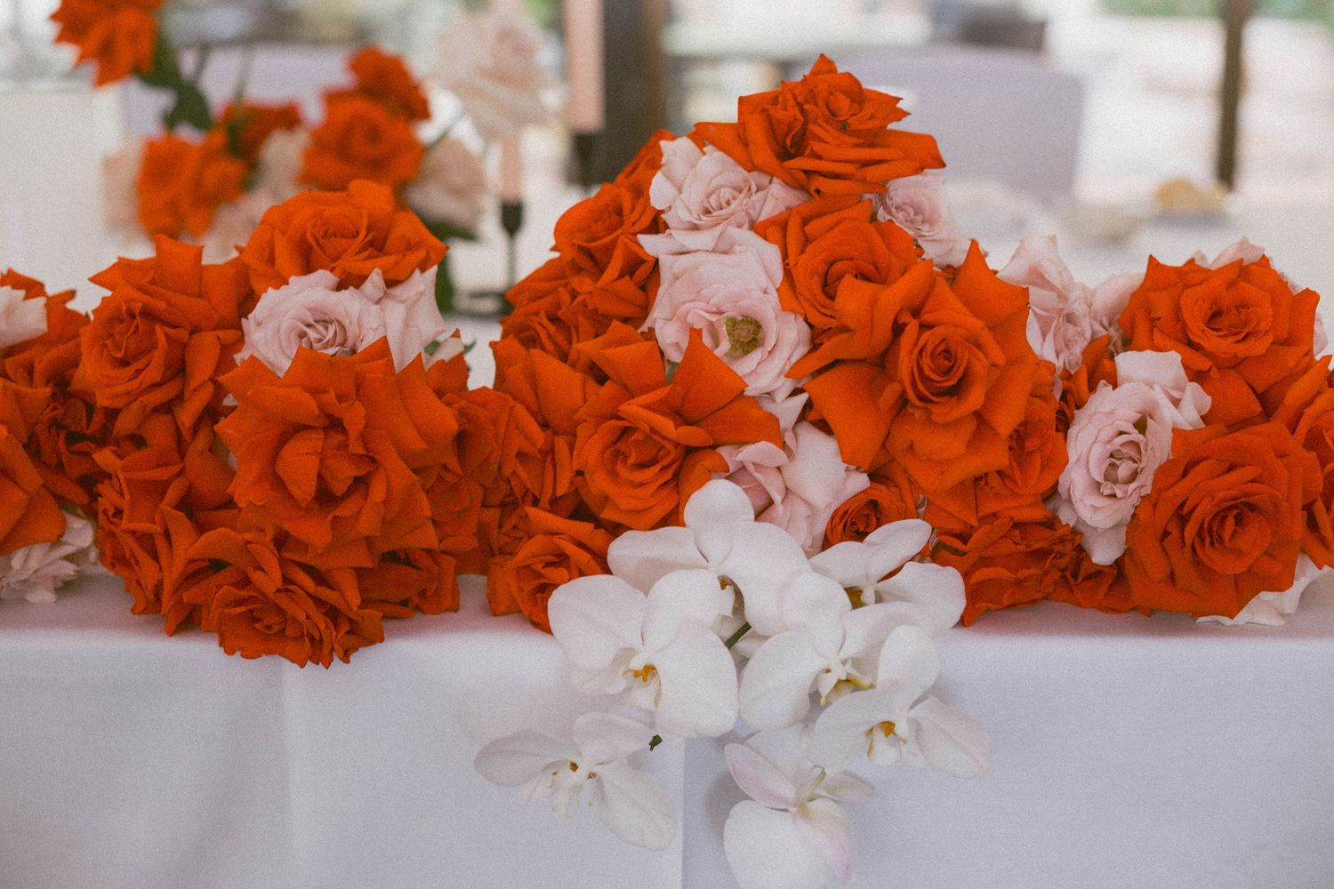 A stunning wedding arrangement featuring bright orange roses and white orchids displayed on a white tablecloth.