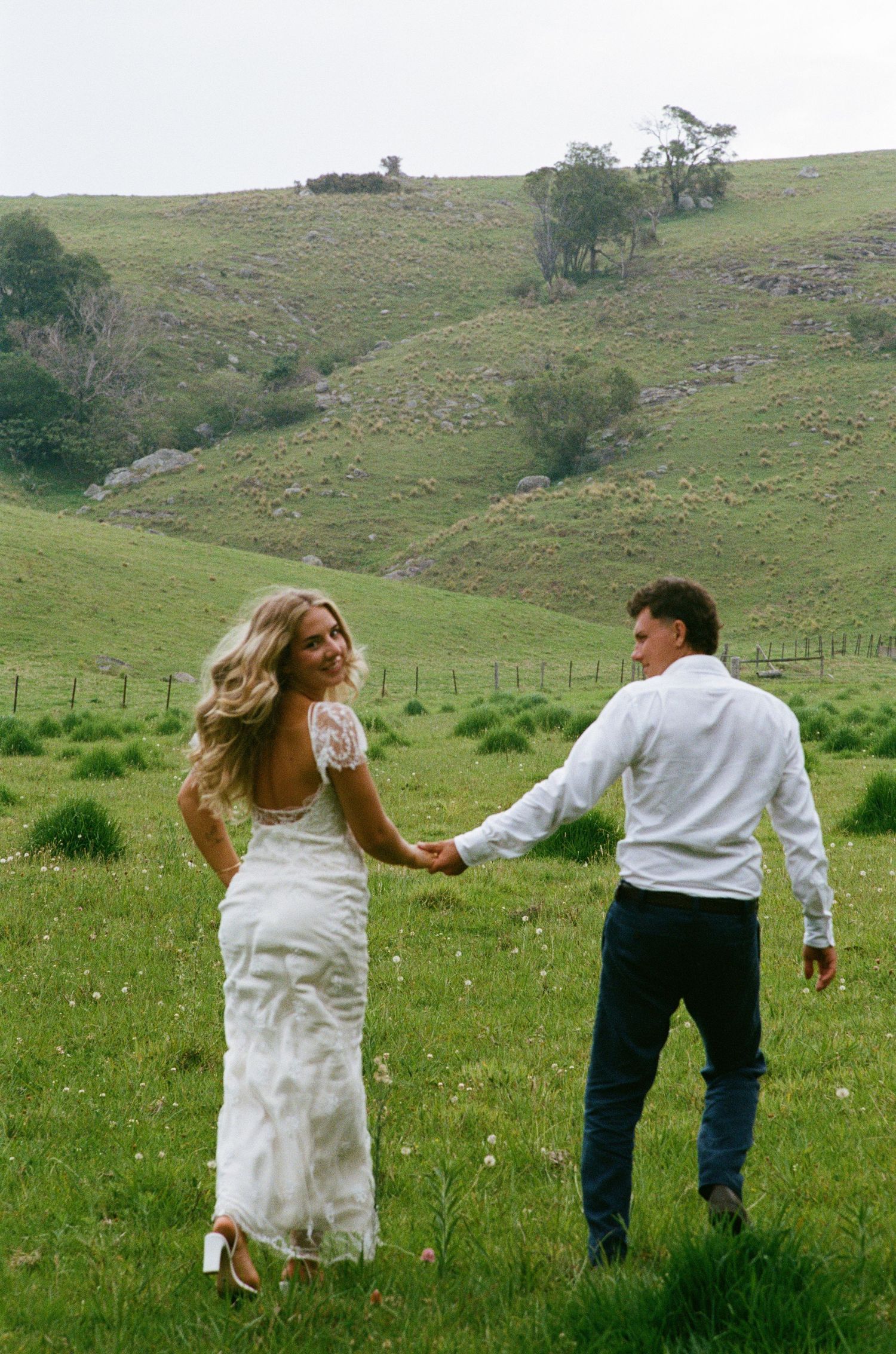 Couple dances playfully in a green meadow with rolling hills behind them.