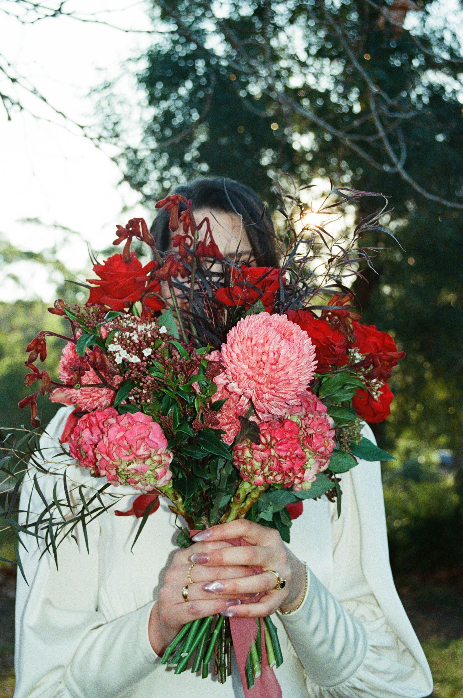 A beautiful bouquet of pink dahlias and red flowers held against a white sweater with a dreamy outdoor backdrop.