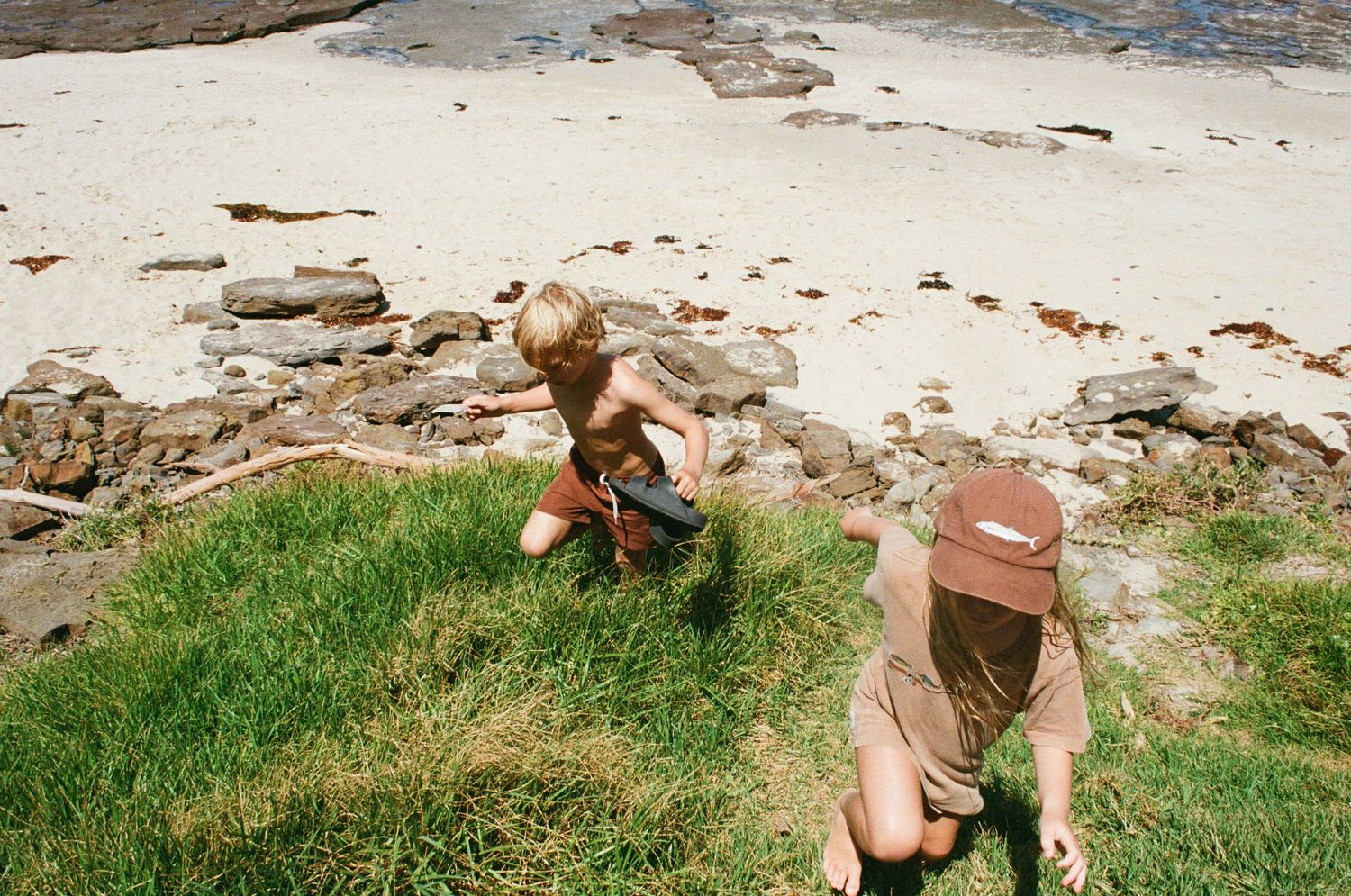 Two people sit on grassy patch near rocky beach area on sunny day.