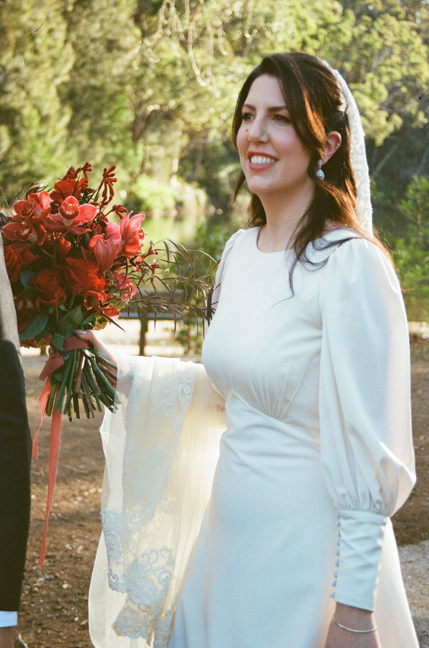 A figure in a white dress holding a bouquet of red roses poses outdoors in a sunlit garden setting.