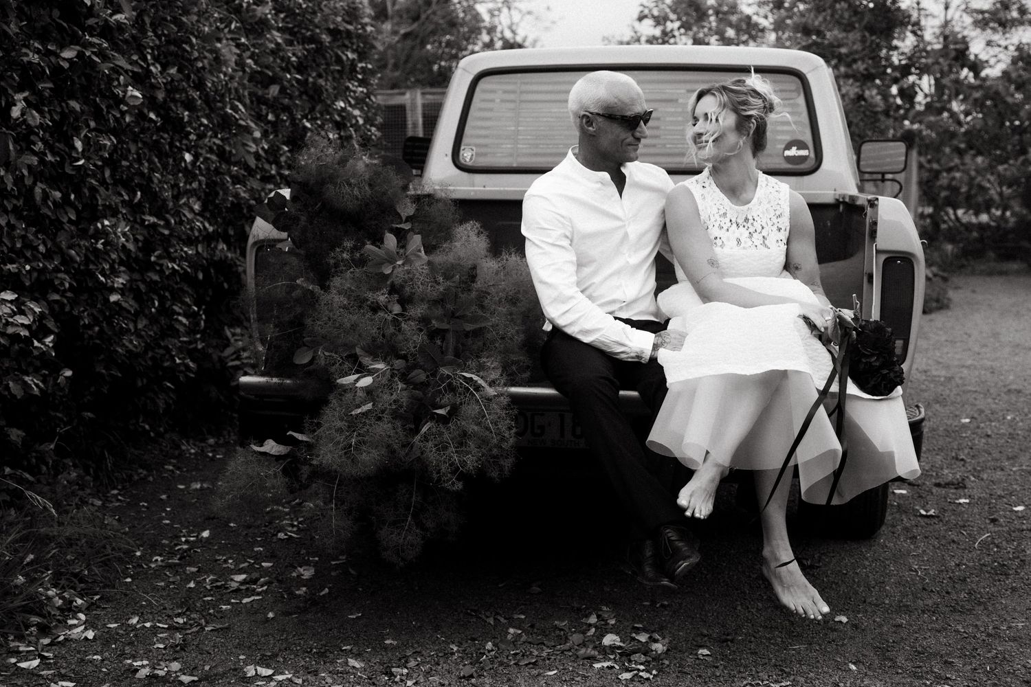 Wedding party gathered around a vintage car outdoors under shady trees.