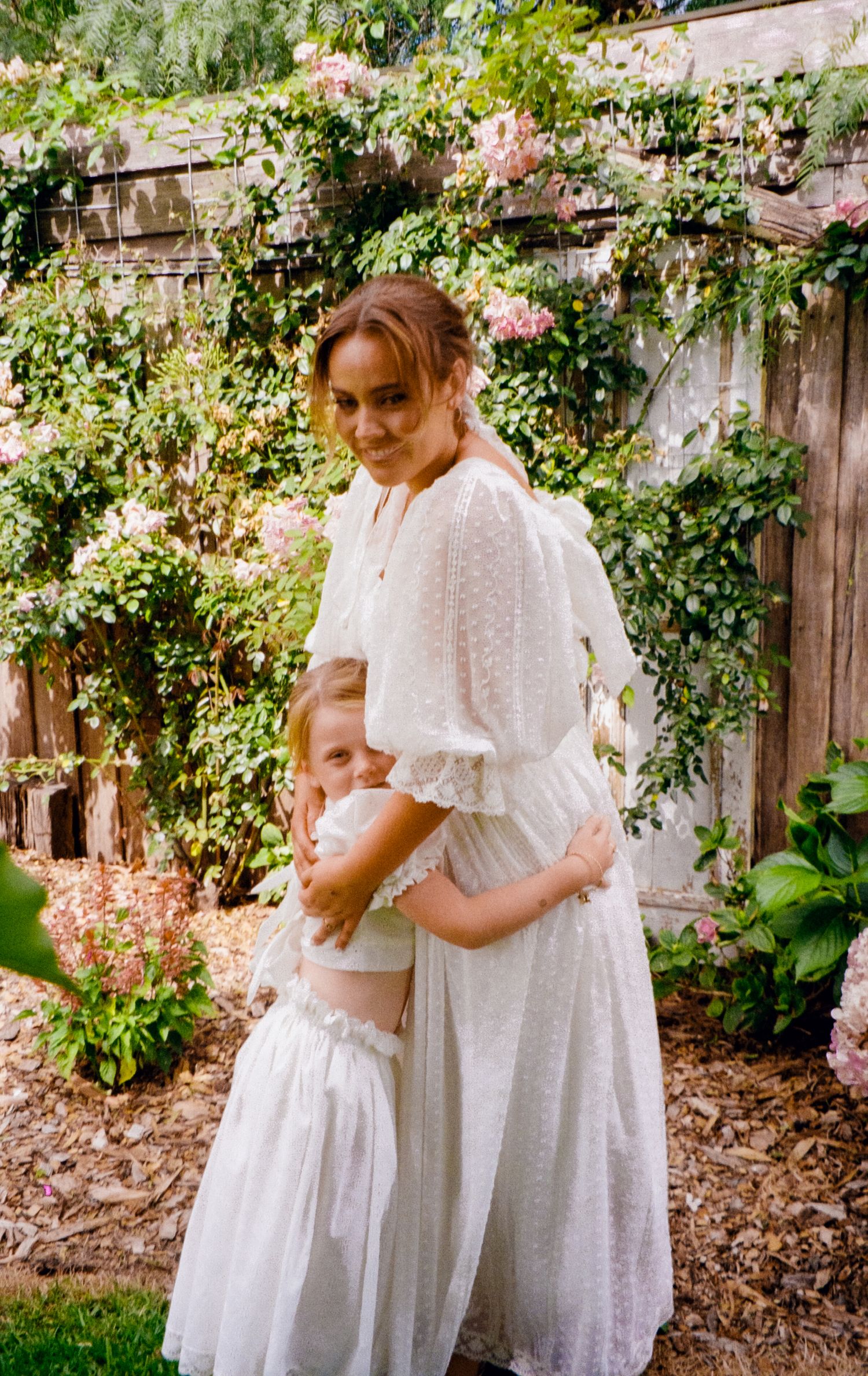 A heartwarming moment captured in a garden with climbing roses and wooden fence backdrop.