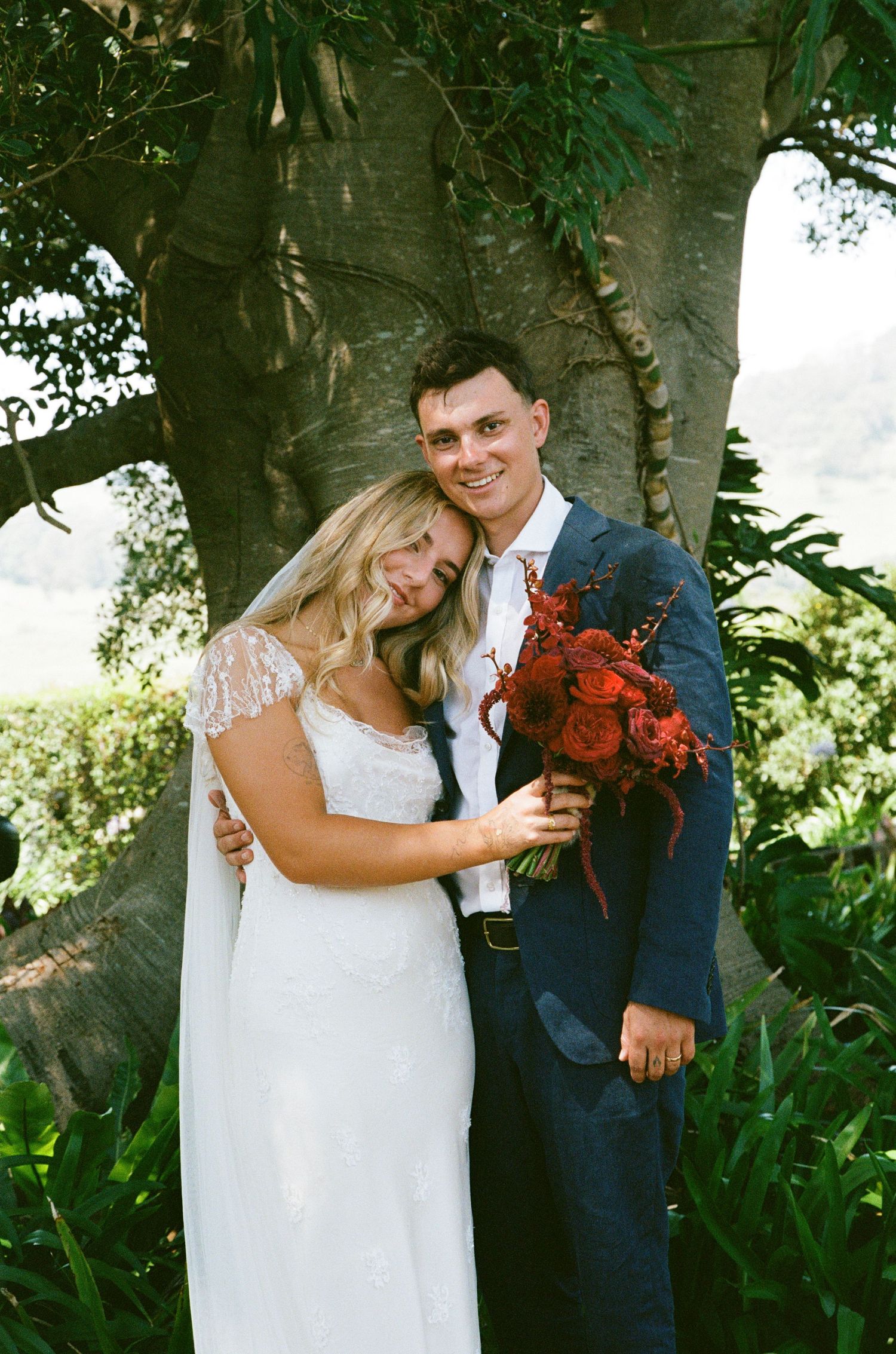 Wedding couple poses with red bouquet against garden greenery.