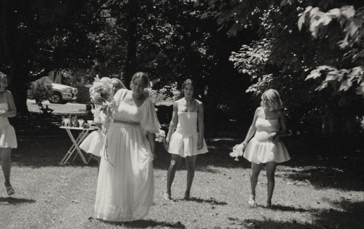 A group wearing white formal dresses poses outdoors near trees at an elegant garden event.
