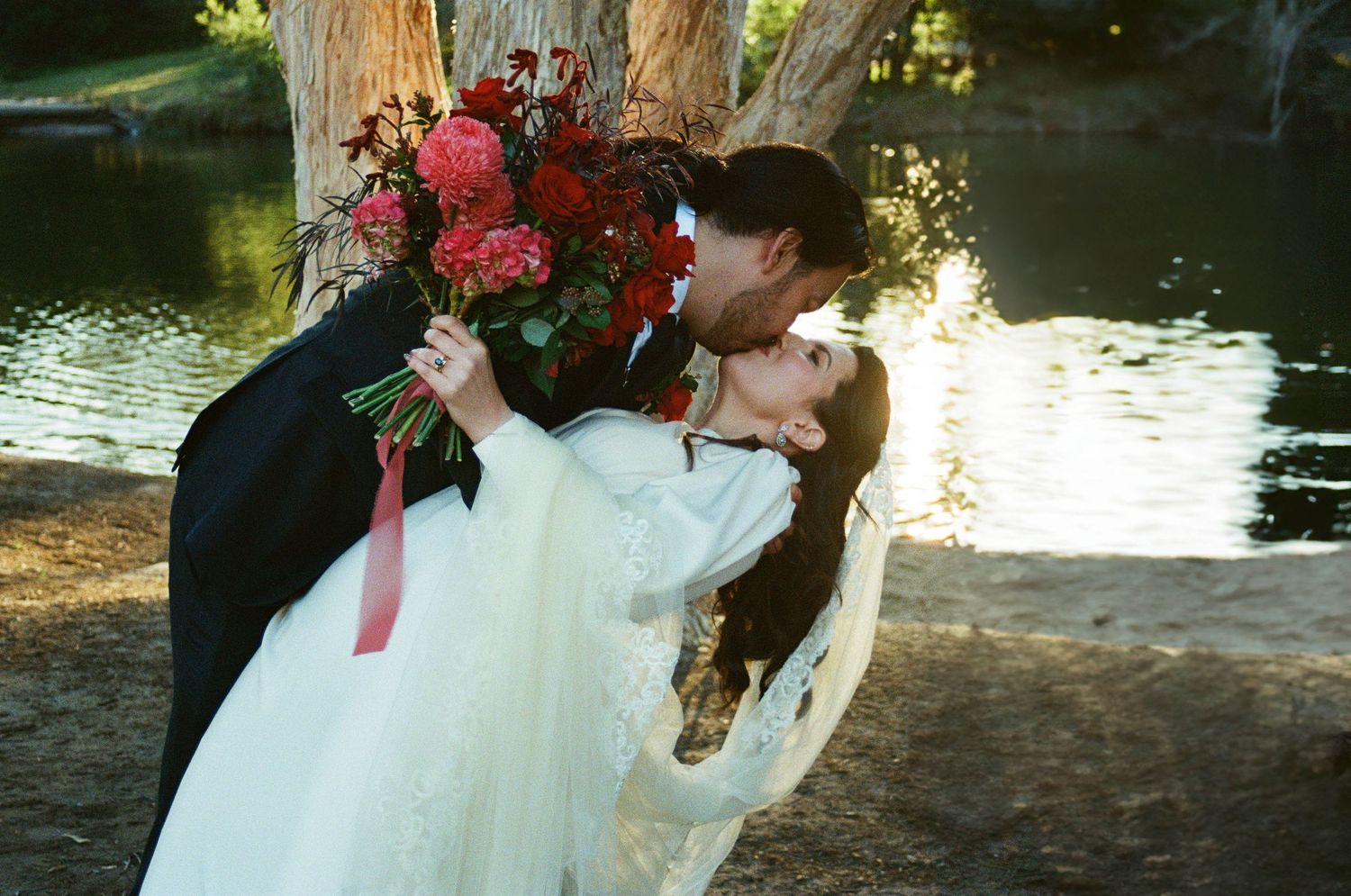 A romantic outdoor wedding scene with red floral bouquet by a pond in natural sunlight.