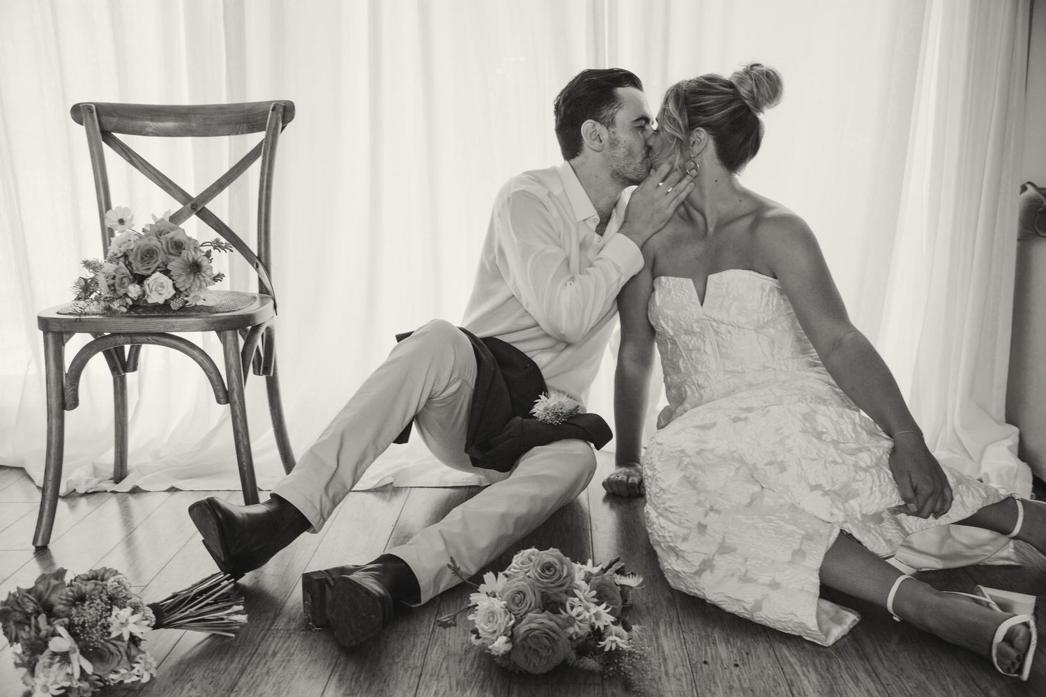 Black and white photo of a romantic moment between newlyweds sitting on floor with vintage chair and floral details.