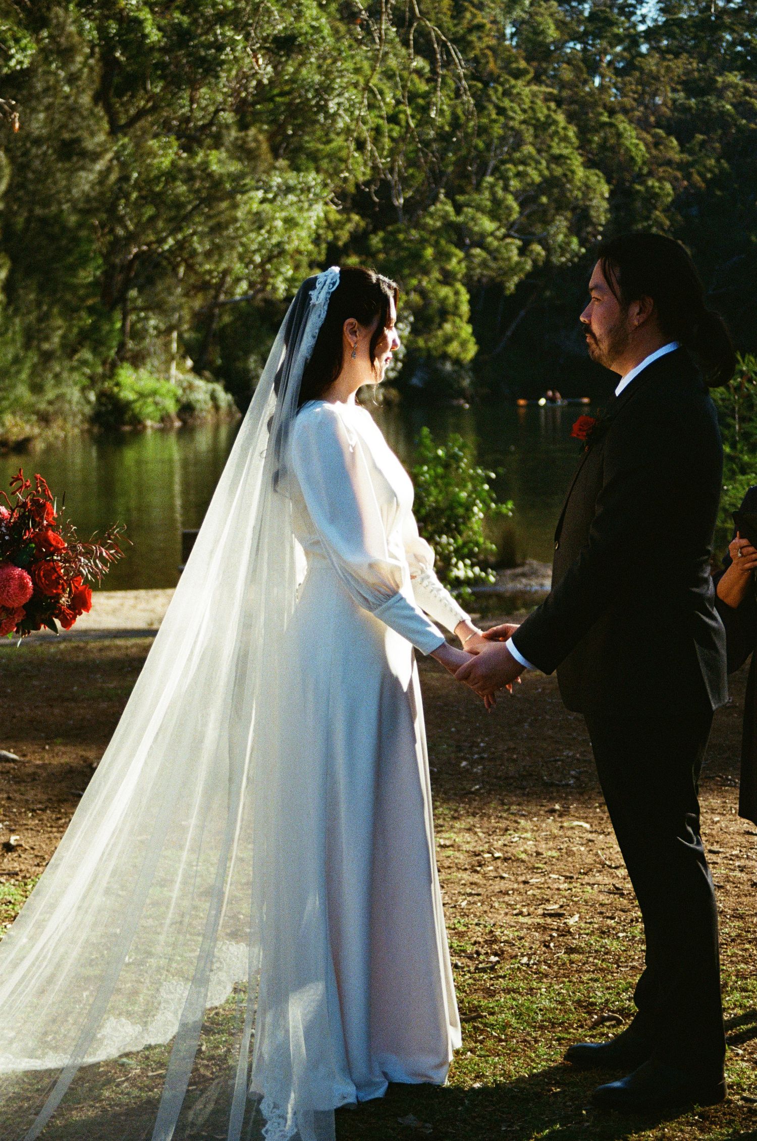 A wedding ceremony moment by a pond with greenery and red floral decorations in the background.