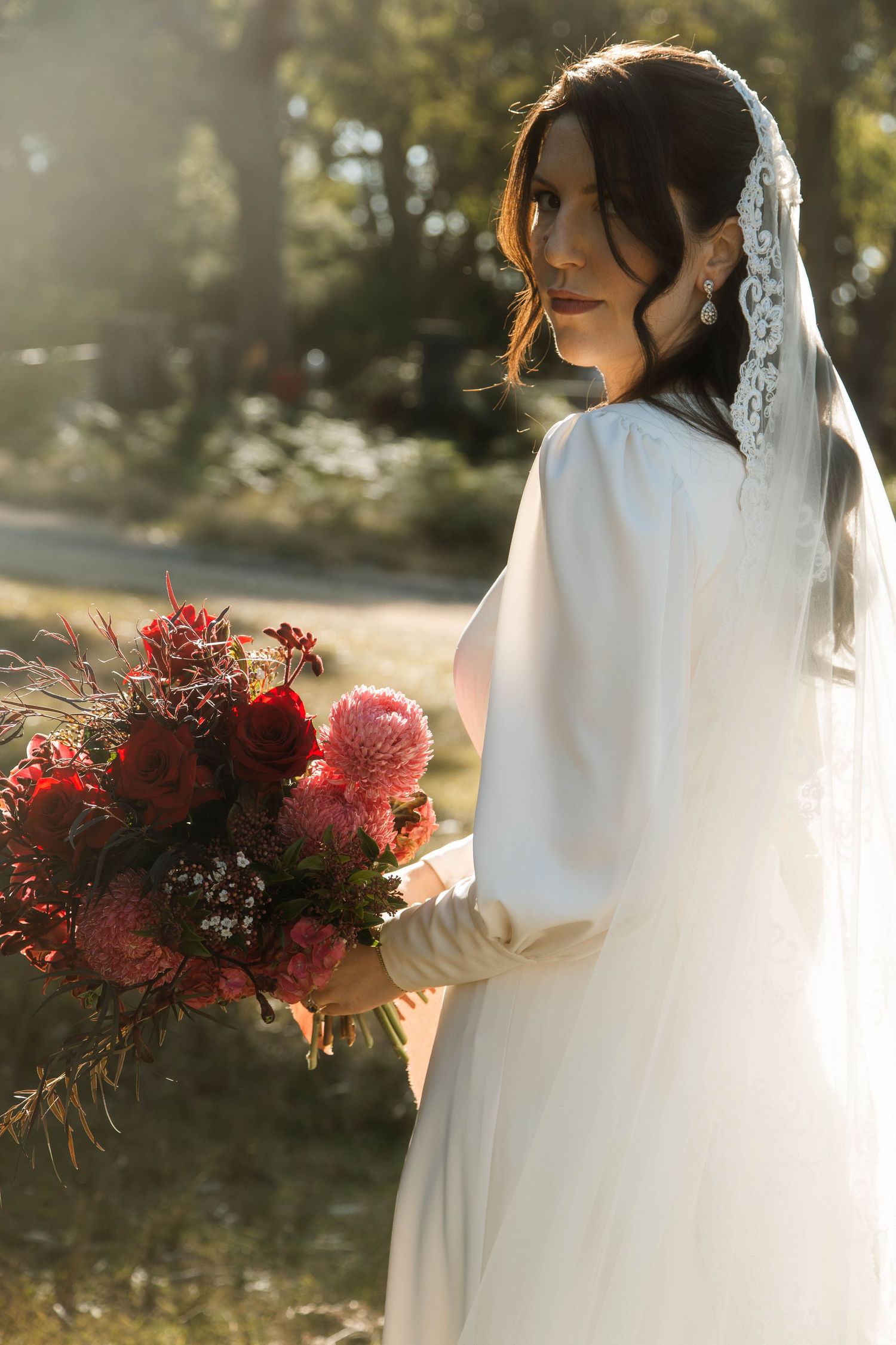 Bride in elegant white dress holds vibrant red and pink bouquet in golden sunlit garden setting.
