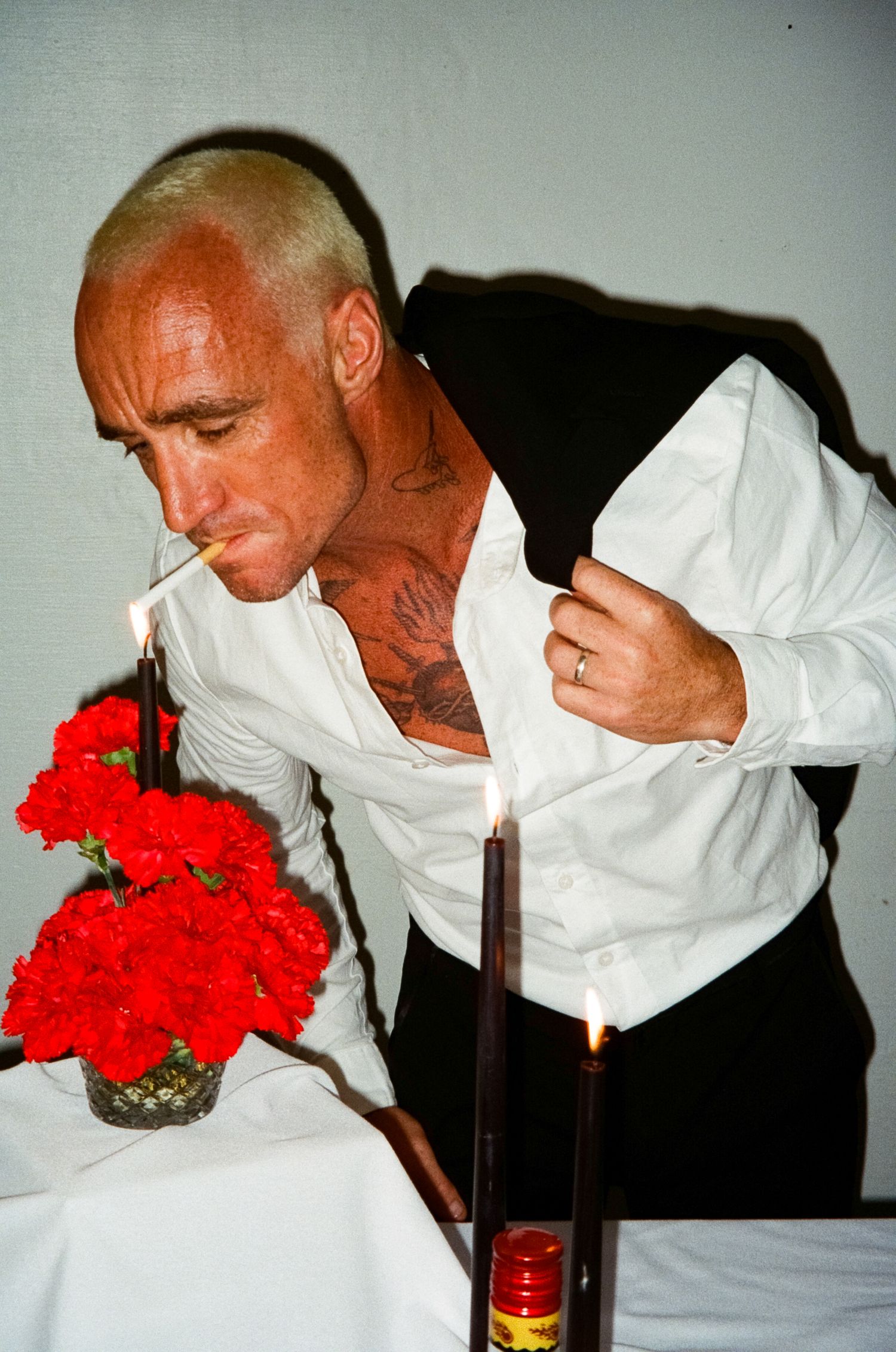 A figure in white shirt smells red carnations on a table with a candle and bottle.