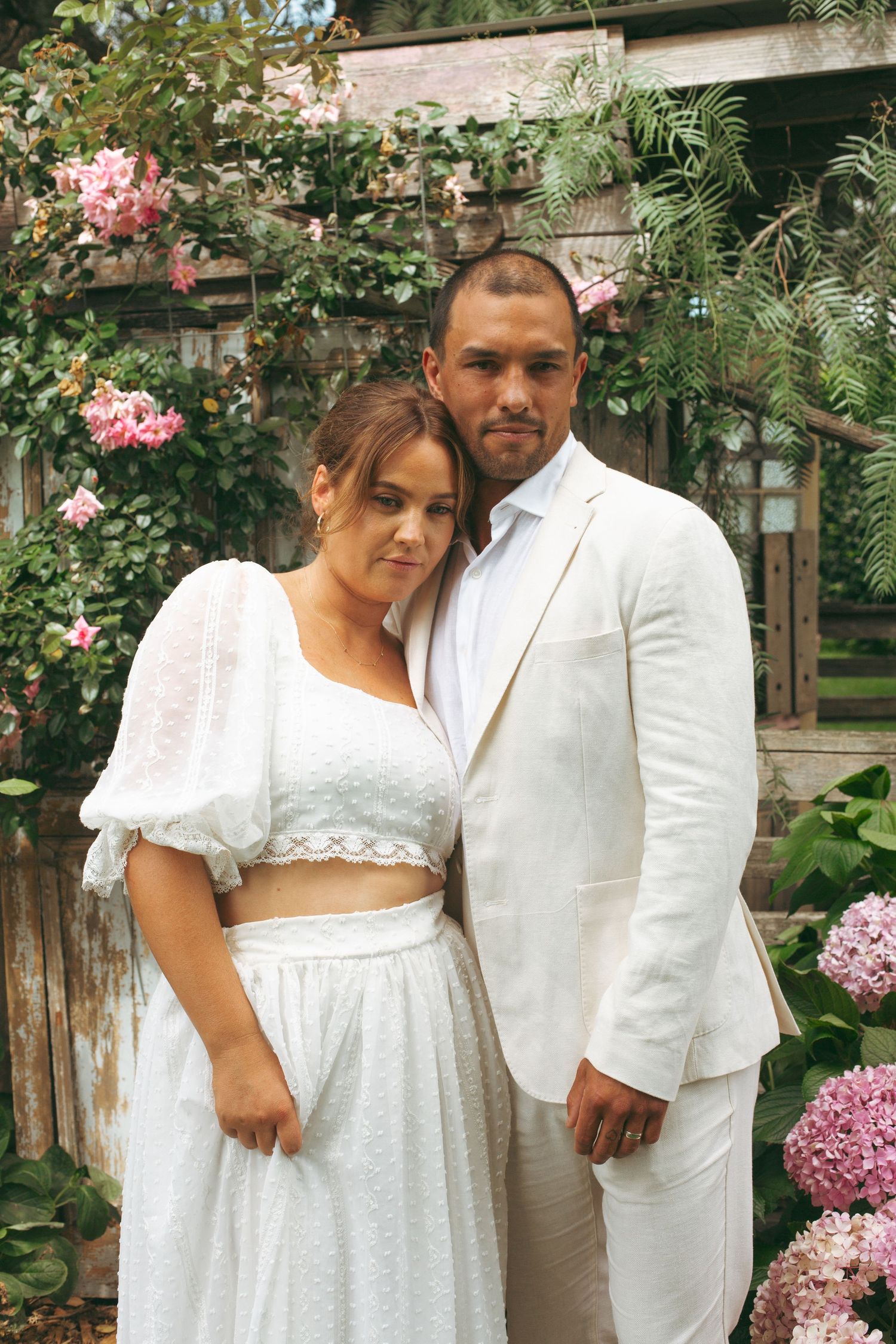 Wedding couple poses in white attire against a romantic garden backdrop with blooming pink flowers.