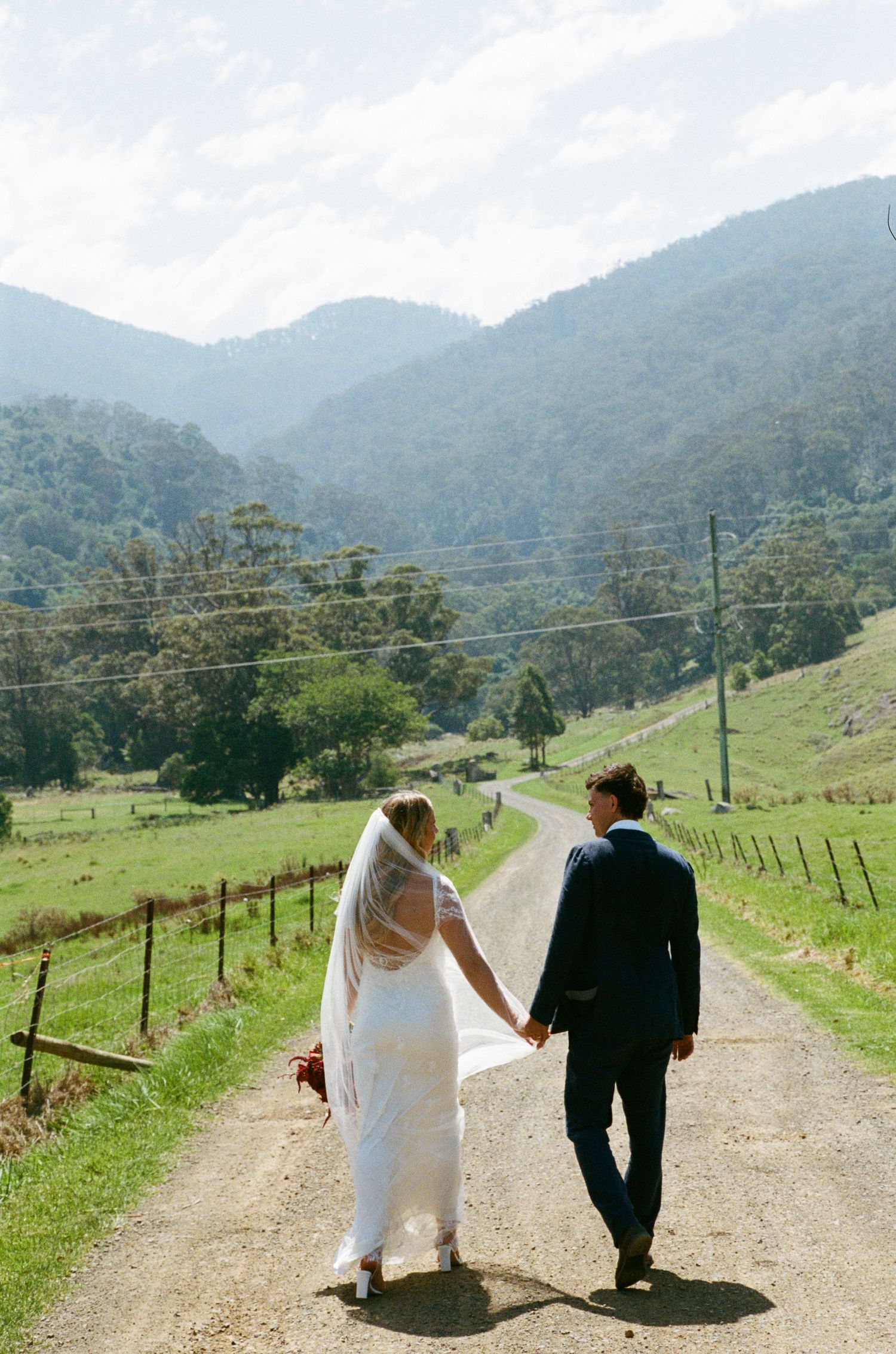 A couple walks down a country road with mountains in the background.