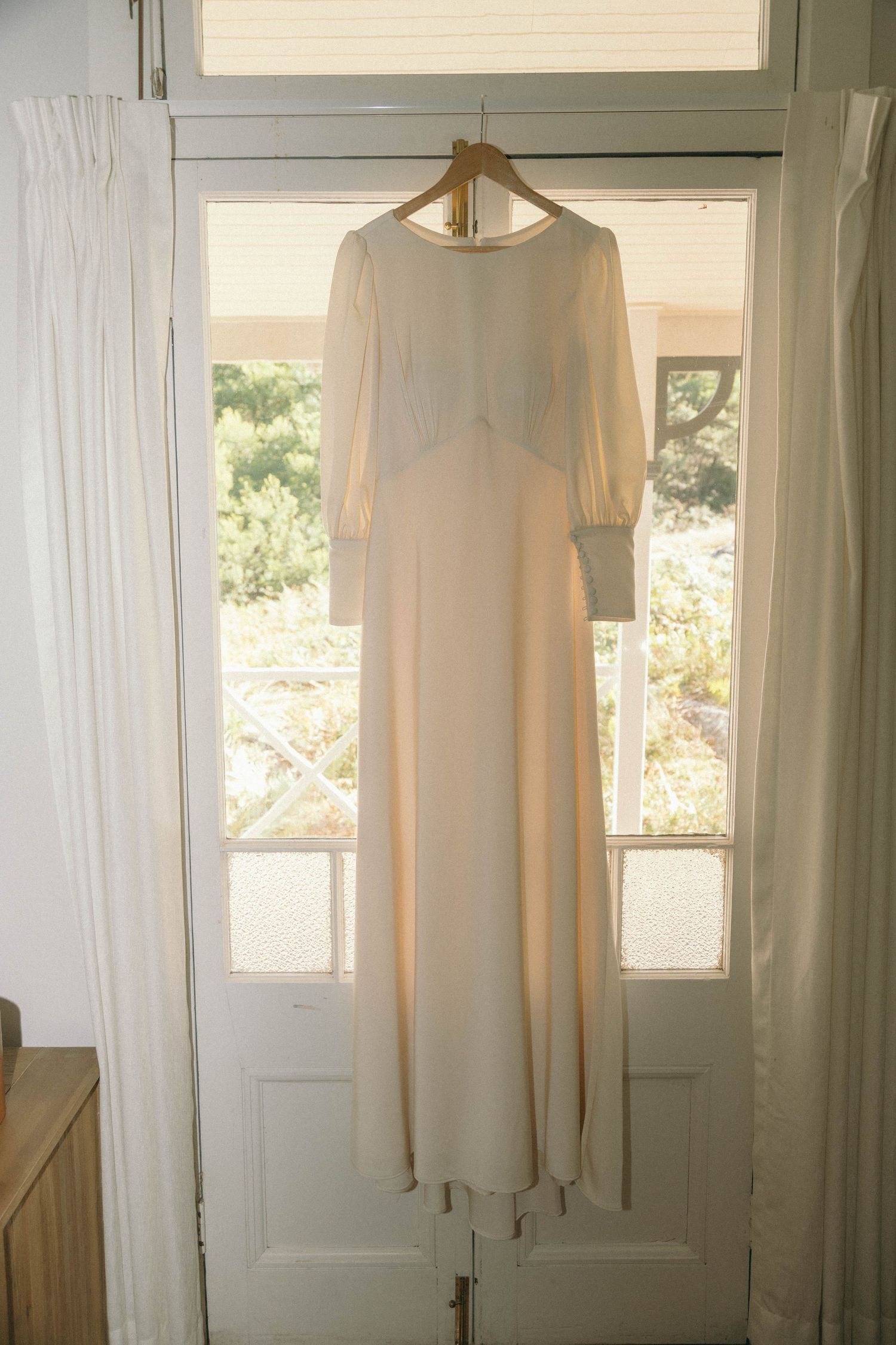 Sunlit wedding dress hanging in a bright window with sheer white curtains.