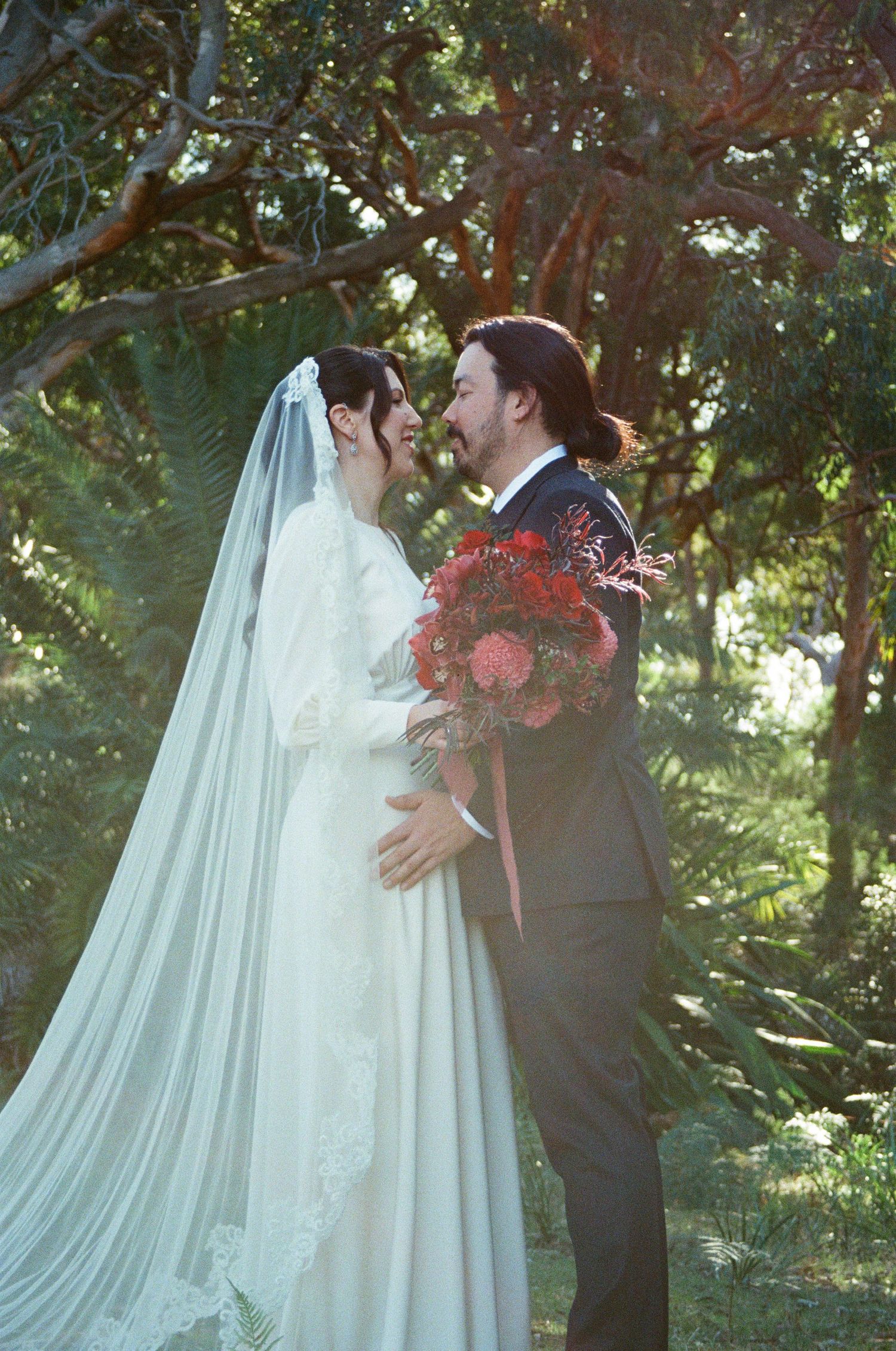 A romantic wedding couple shares intimate moments in a lush garden setting with flowing white veil and red bouquet.