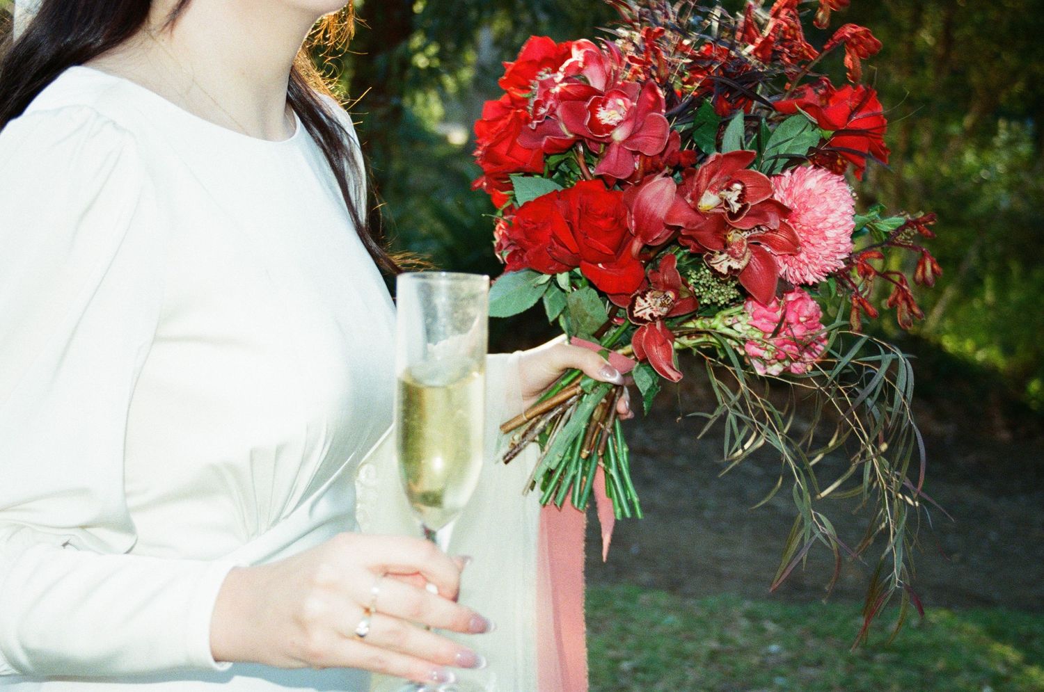 Close-up of wedding bouquet with vibrant red and pink flowers against greenery in outdoor garden setting.