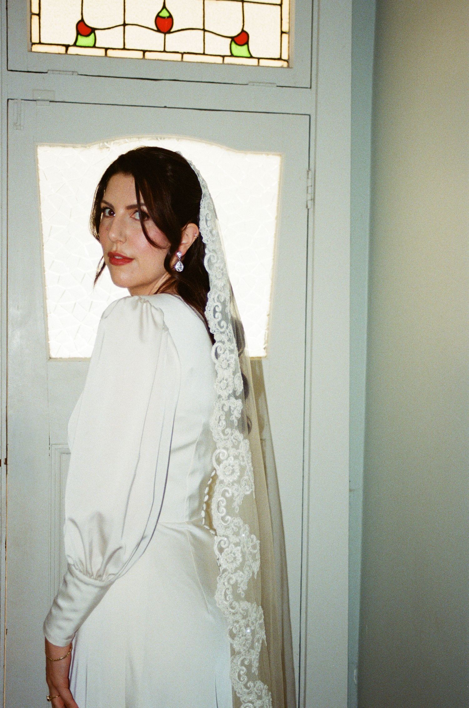 A bride in an elegant long-sleeved white gown with lace veil poses by a stained glass window.