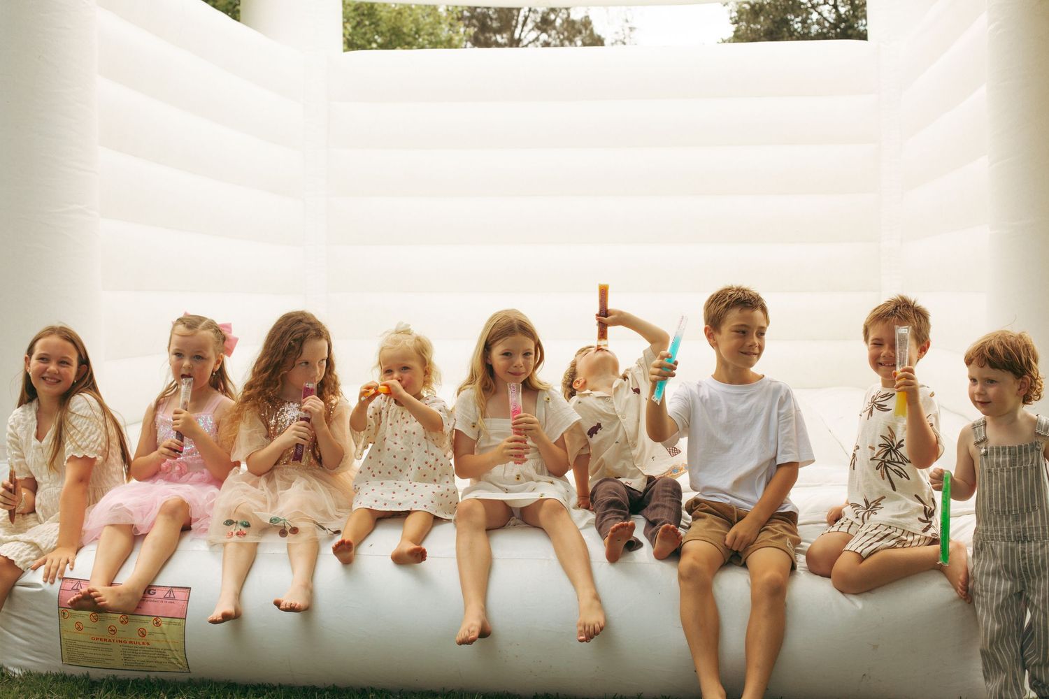 Children sitting in a row on an inflatable surface eating popsicles and ice cream treats on a sunny day.