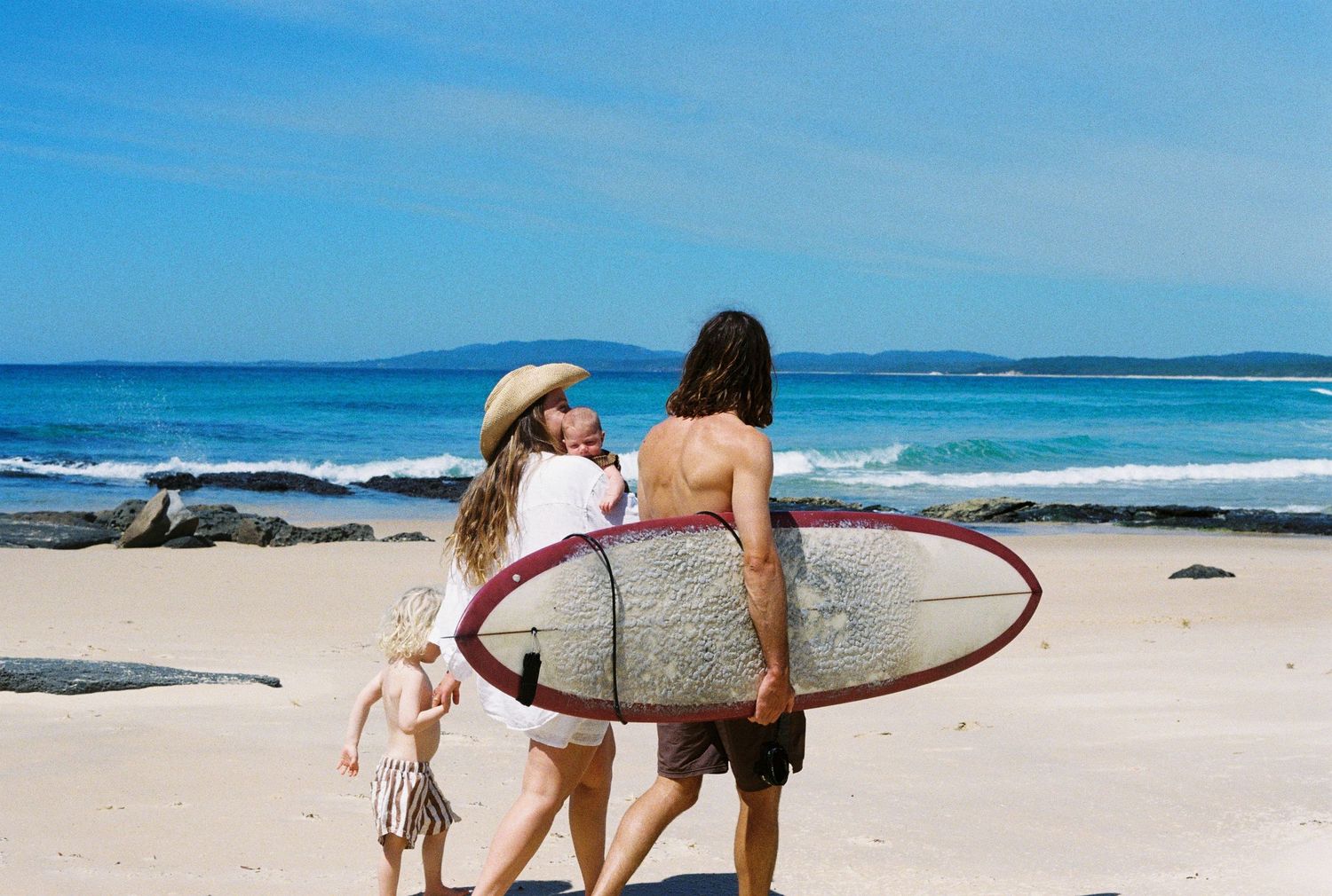 Surfers walking along a scenic beach with surfboards under blue skies and rolling waves.