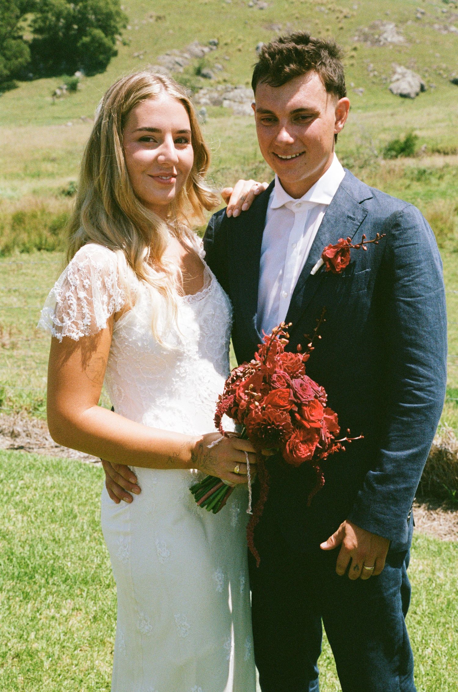 Newlyweds share joyful moments with red floral bouquet in outdoor setting.