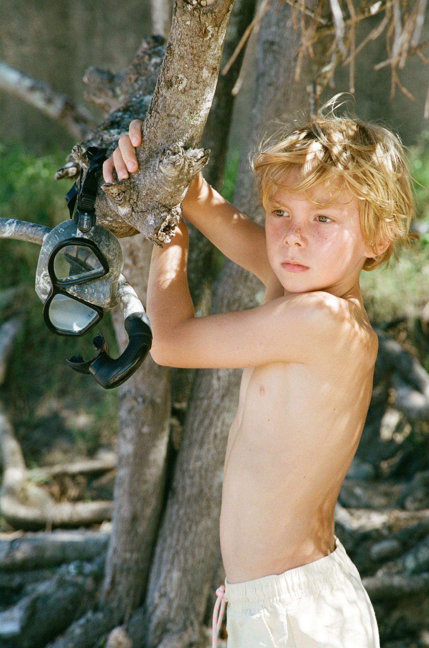 A child in white shorts climbs a tree trunk while holding a black object on a sunny day.