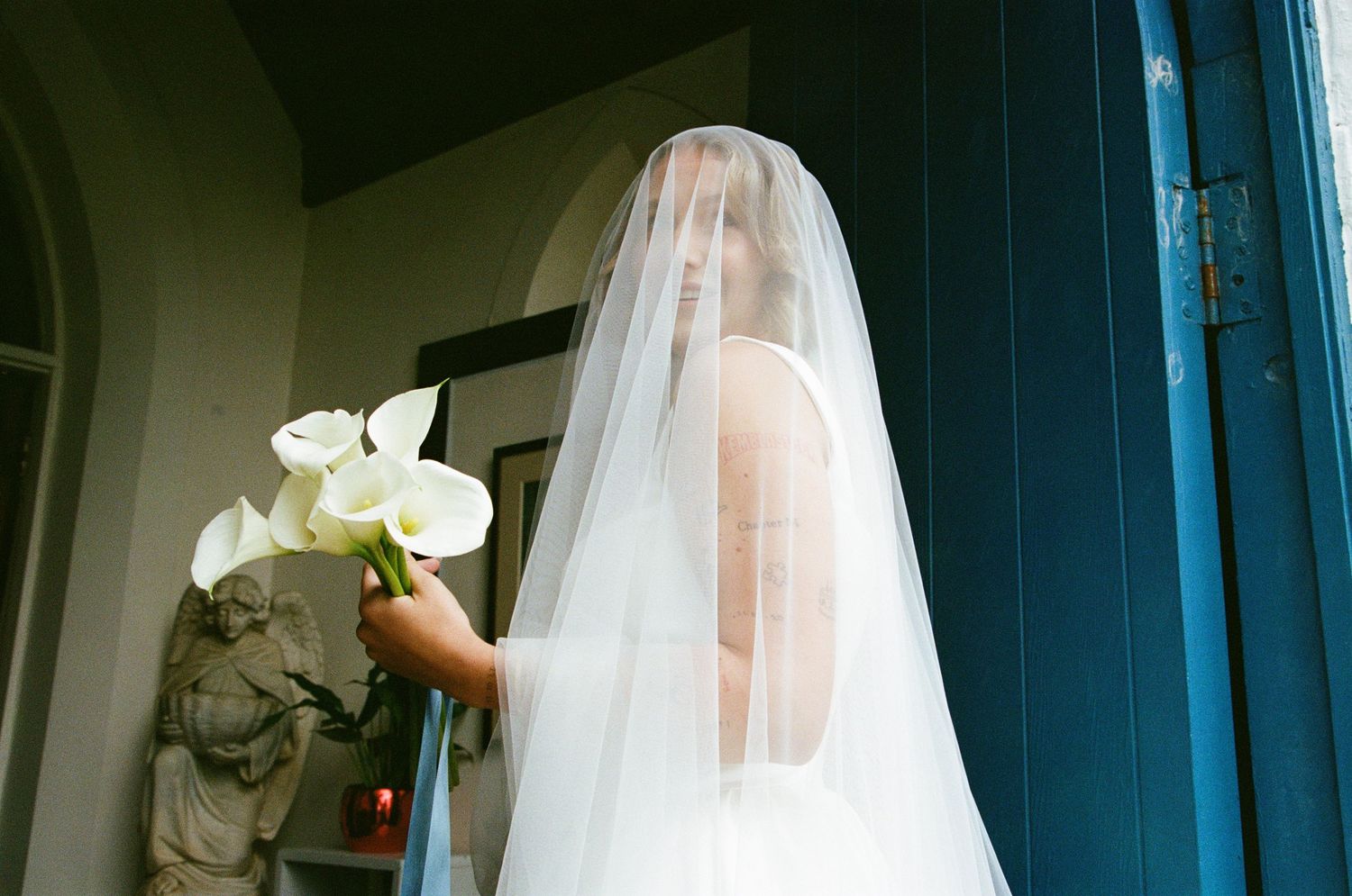 A bride in a flowing white veil stands in front of a blue door during a wedding photoshoot.