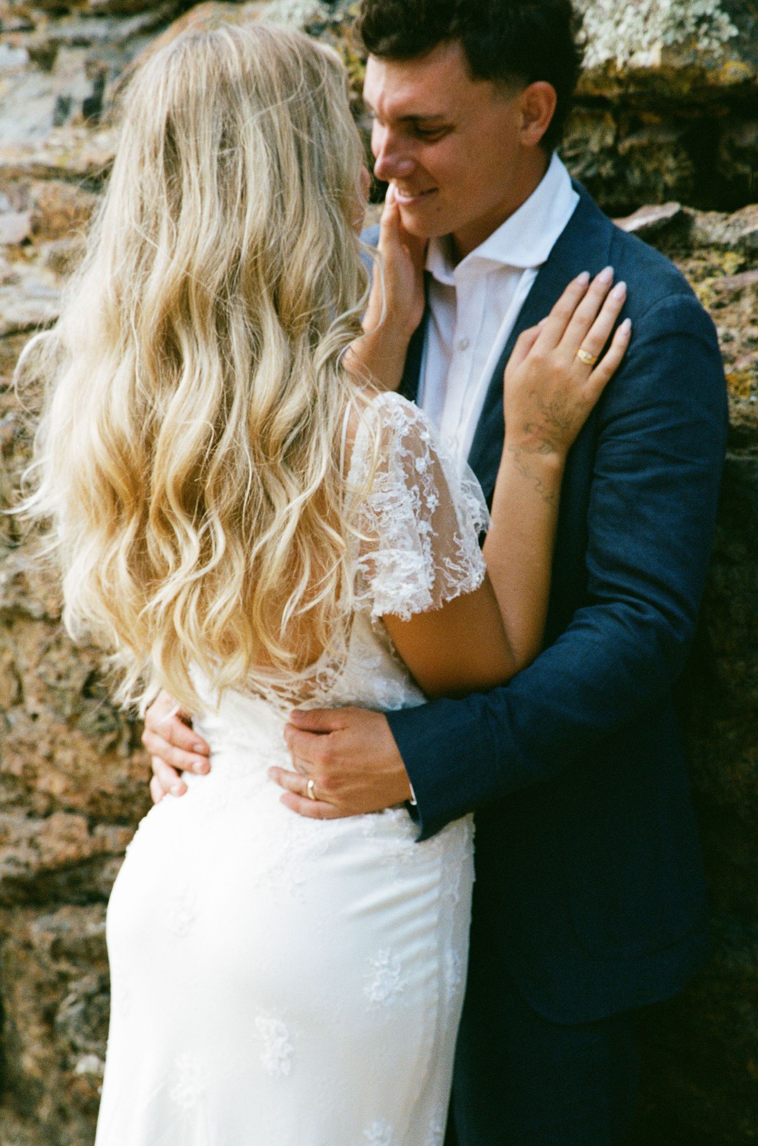 A romantic wedding couple embraces against a rustic stone wall backdrop.