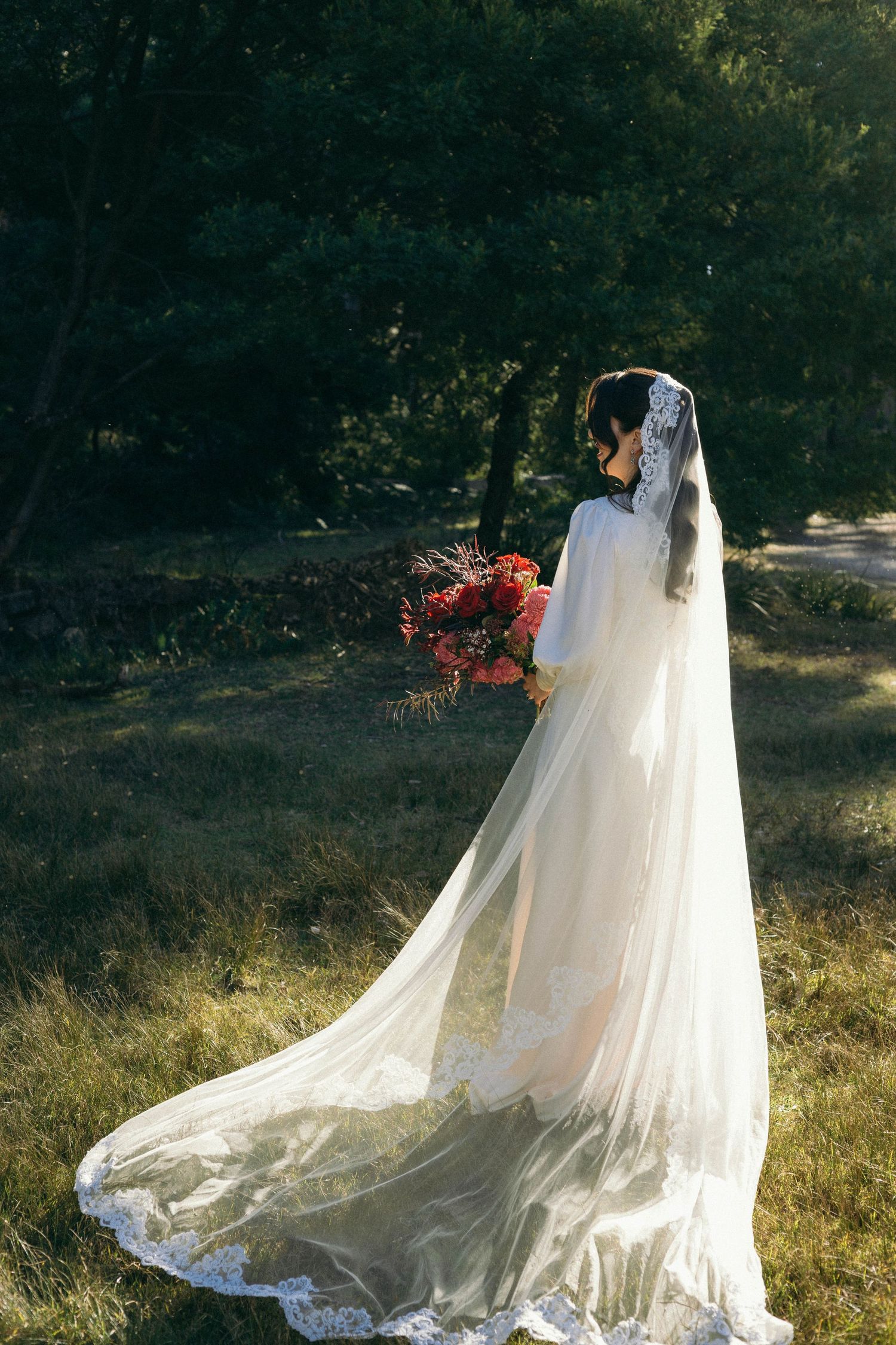 A bride in a flowing white lace gown with long veil holds red flowers while standing in a sunlit garden setting.
