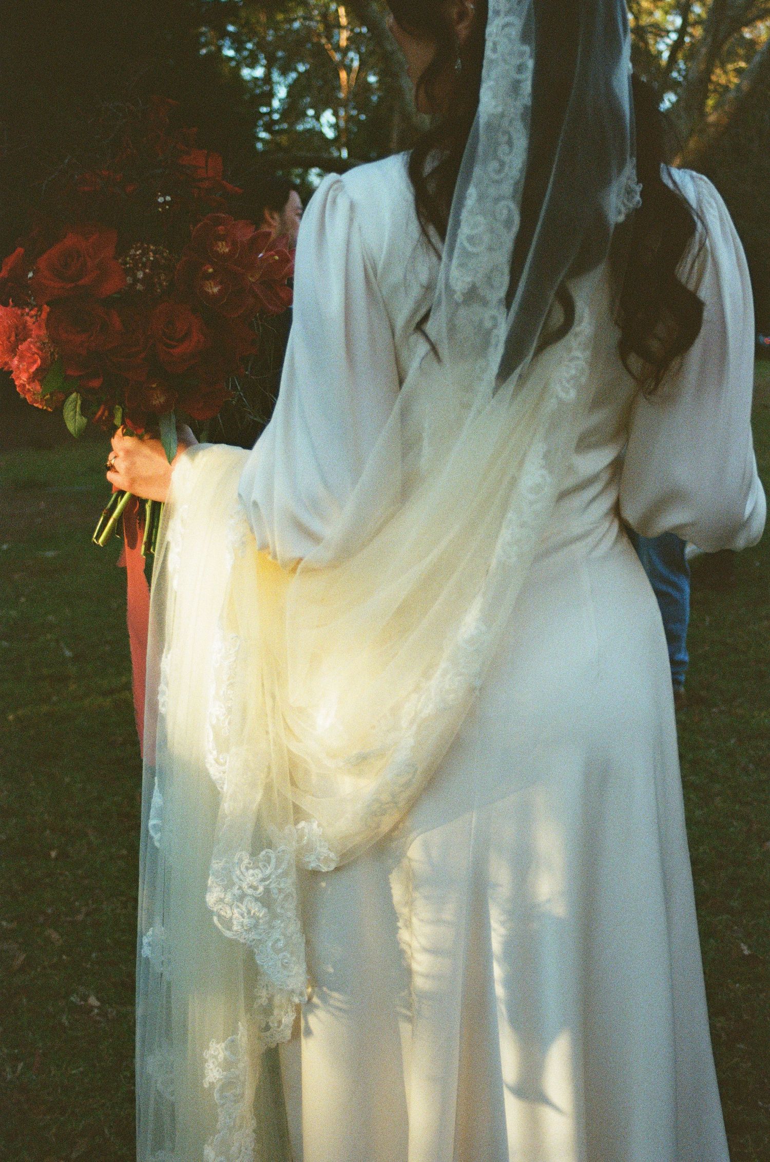 Back view of a bride in a flowing white vintage dress with lace details and long sleeves in a garden setting.