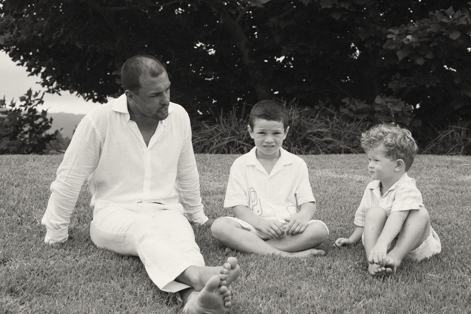 People dressed in white sitting casually on grass under shade trees in a park setting.