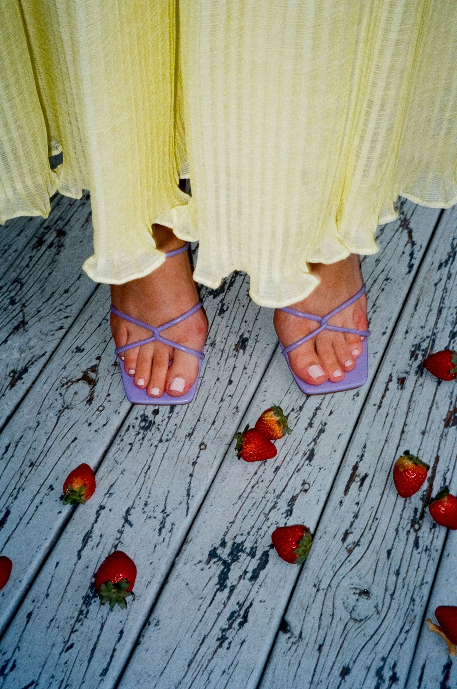 Purple strappy sandals and yellow dress hem on weathered deck with scattered red strawberries.
