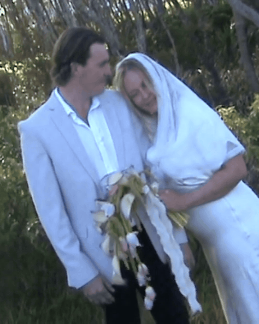 A couple in wedding attire, groom in white suit and bride in white dress with veil, holding a bouquet outdoors.