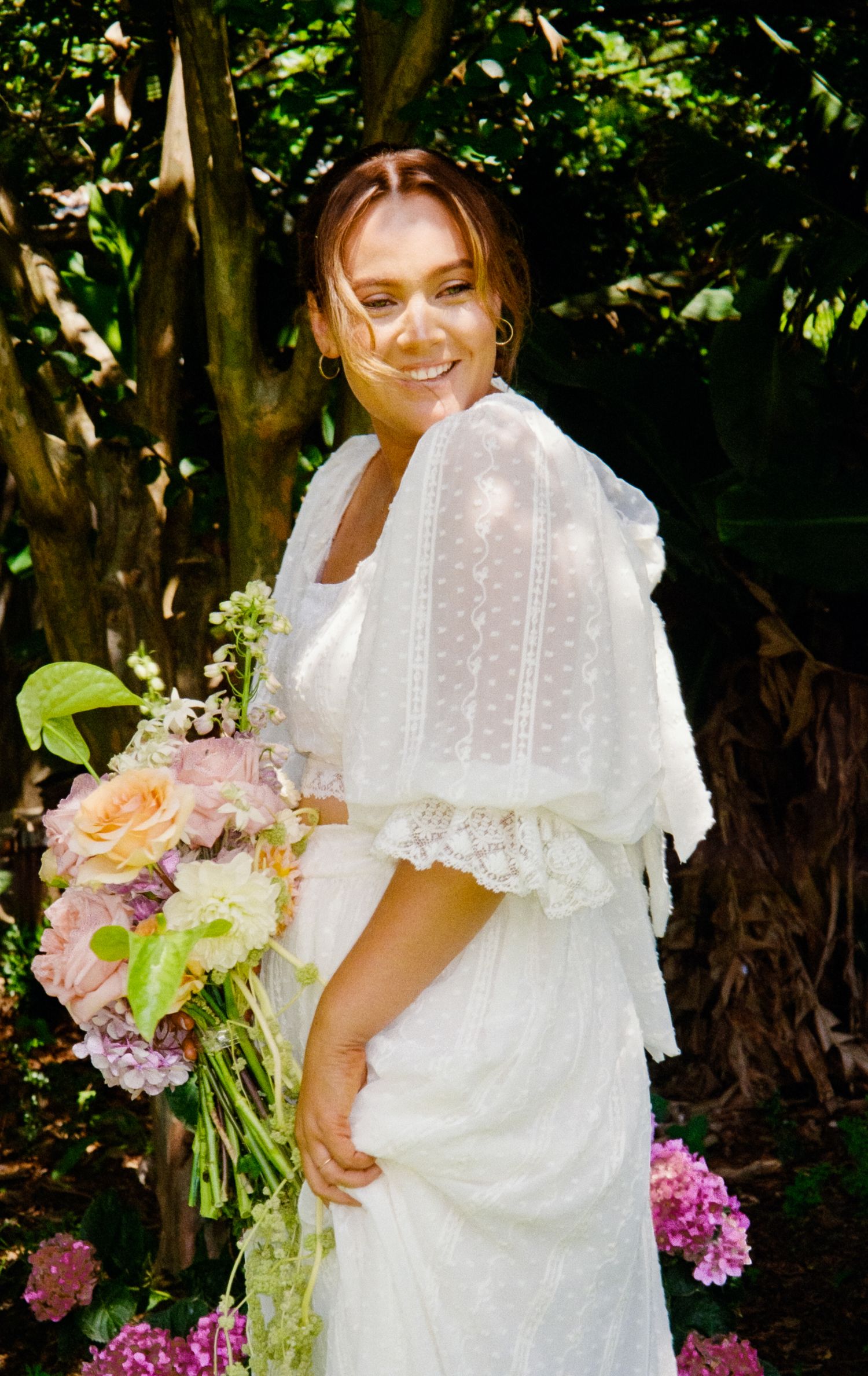 Bride in a vintage-inspired white dress with puff sleeves holds a pastel bouquet in a garden setting.