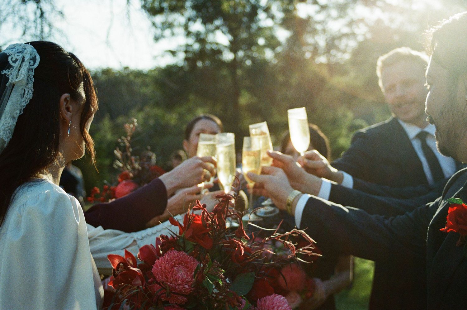 Wedding guests raise champagne glasses during a golden sunset celebration with red floral decorations in view.