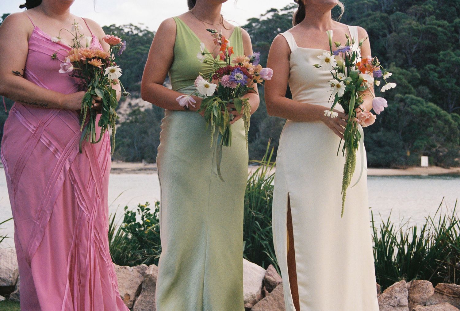Bridesmaids in pink and cream colored dresses holding floral bouquets stand together at an outdoor wedding.