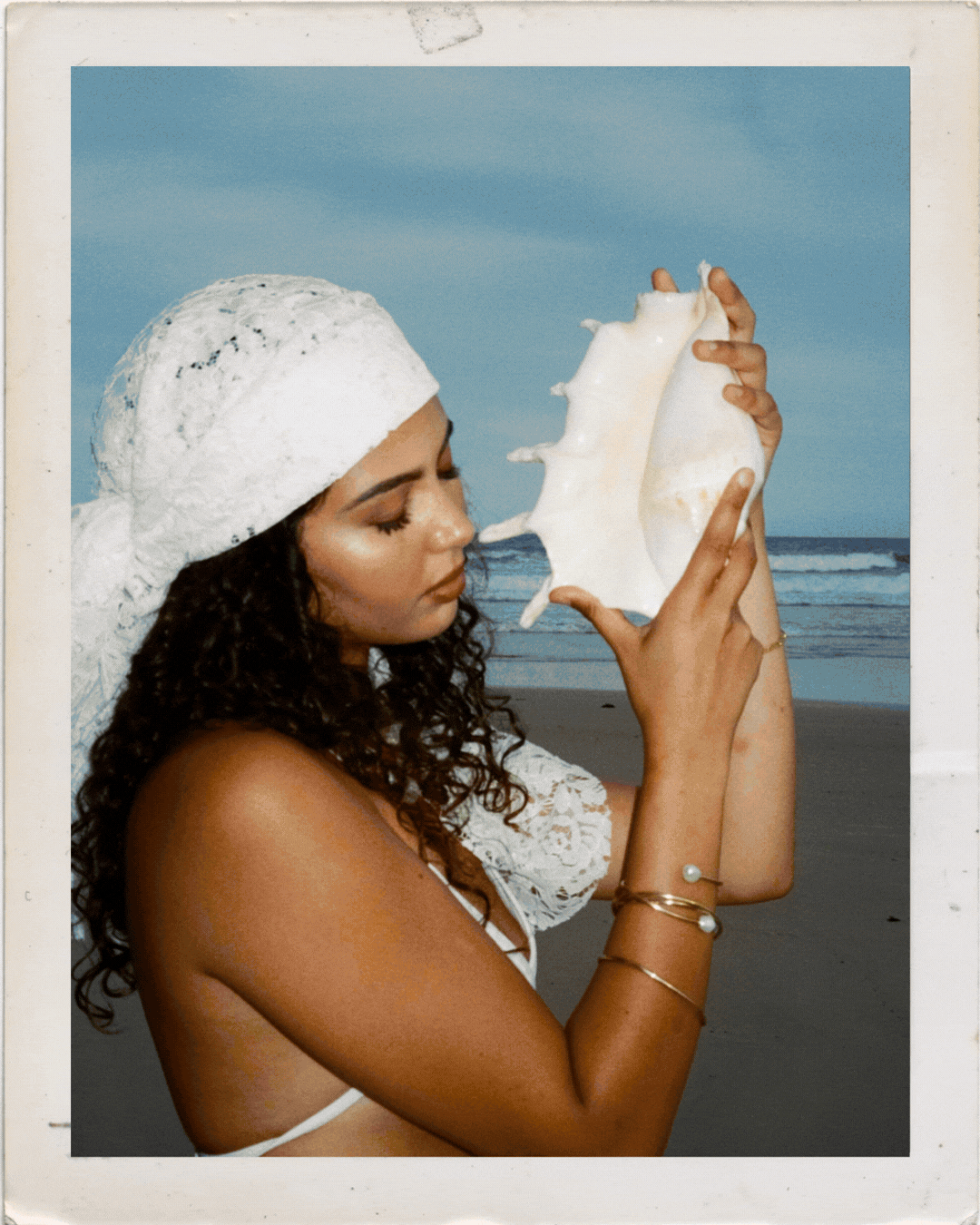 Woman in white crochet hat holds large conch shell at beach, wearing gold bracelets, polaroid style photo.