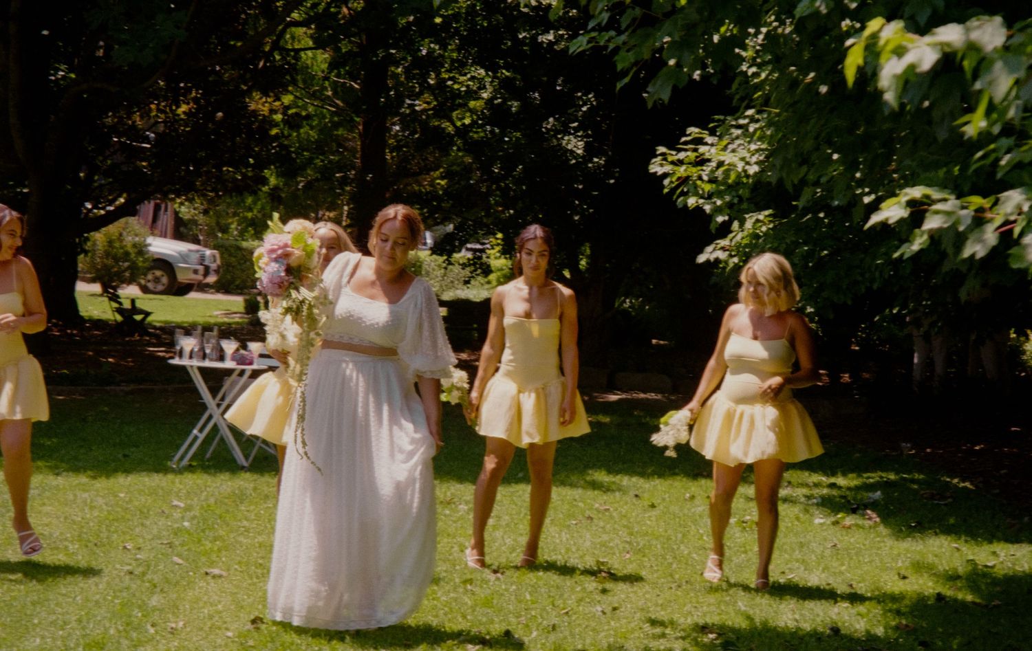 Outdoor summer wedding celebration in a sunlit garden with bridesmaids in yellow dresses and a bride in white.
