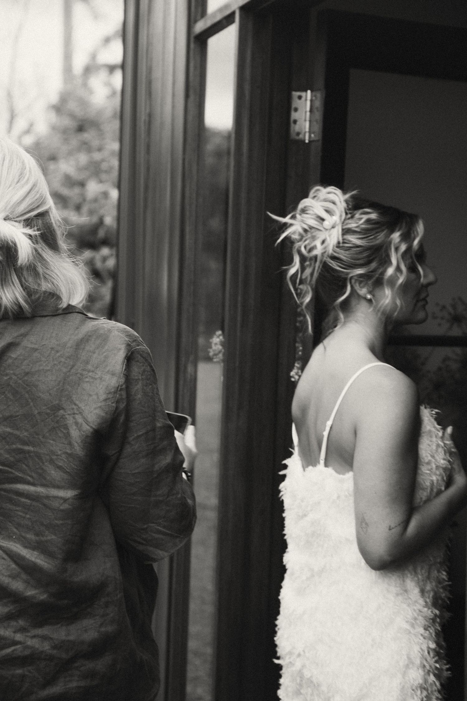 Black and white artistic photo of a spouse in a feathered white dress standing in a doorway.
