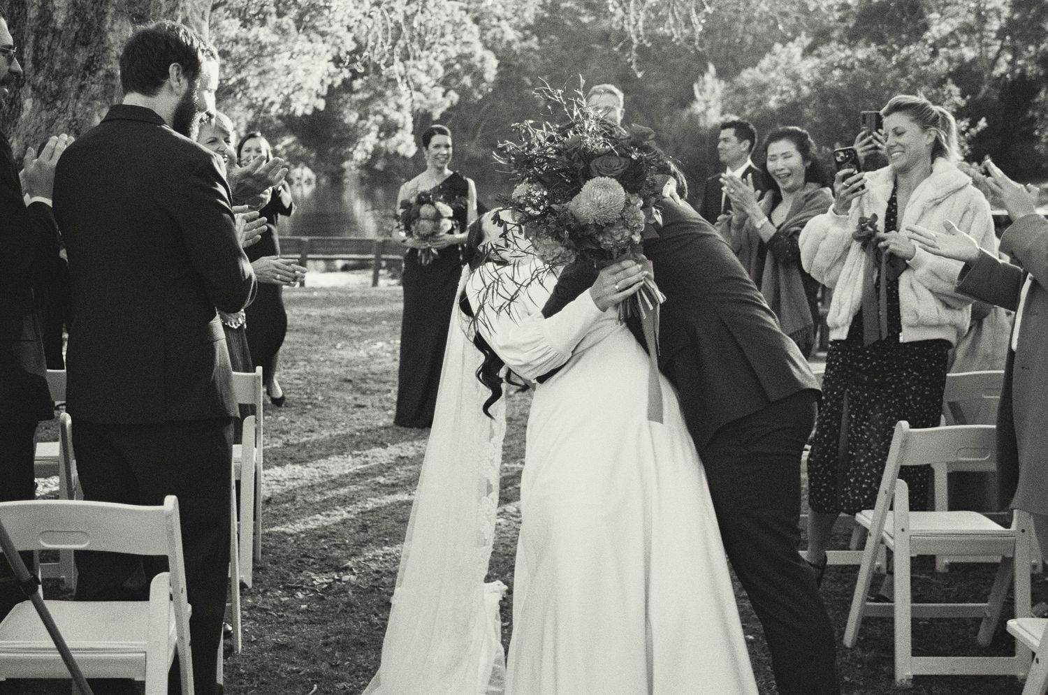Intimate wedding ceremony moment outdoors with guests watching from white folding chairs.