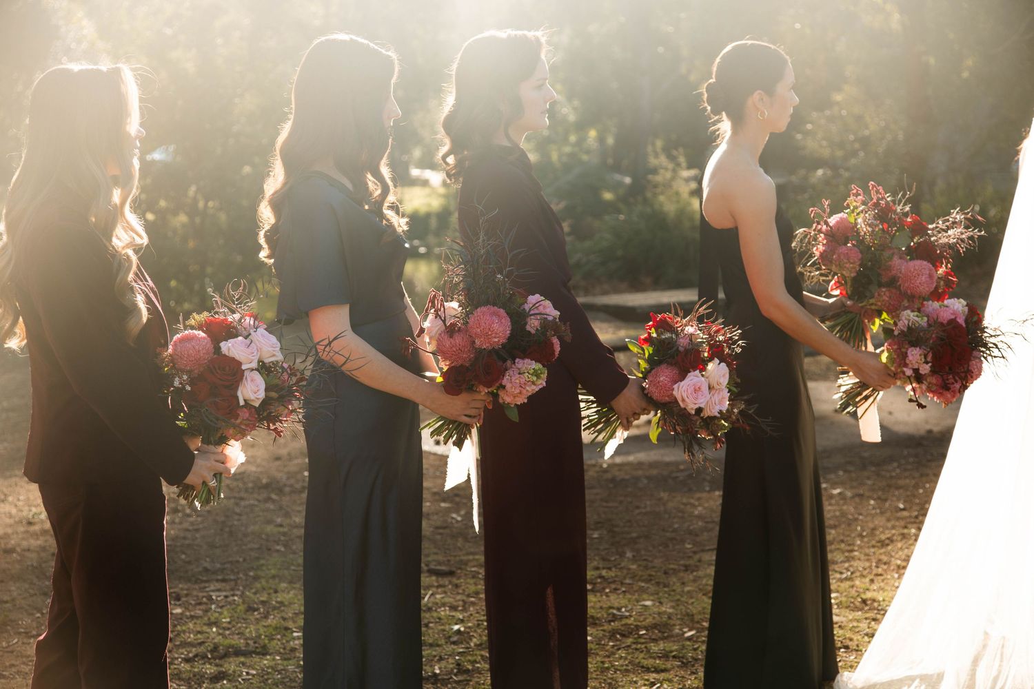 Bridesmaids in elegant black dresses hold colorful bouquets during an outdoor wedding ceremony in golden sunlight.